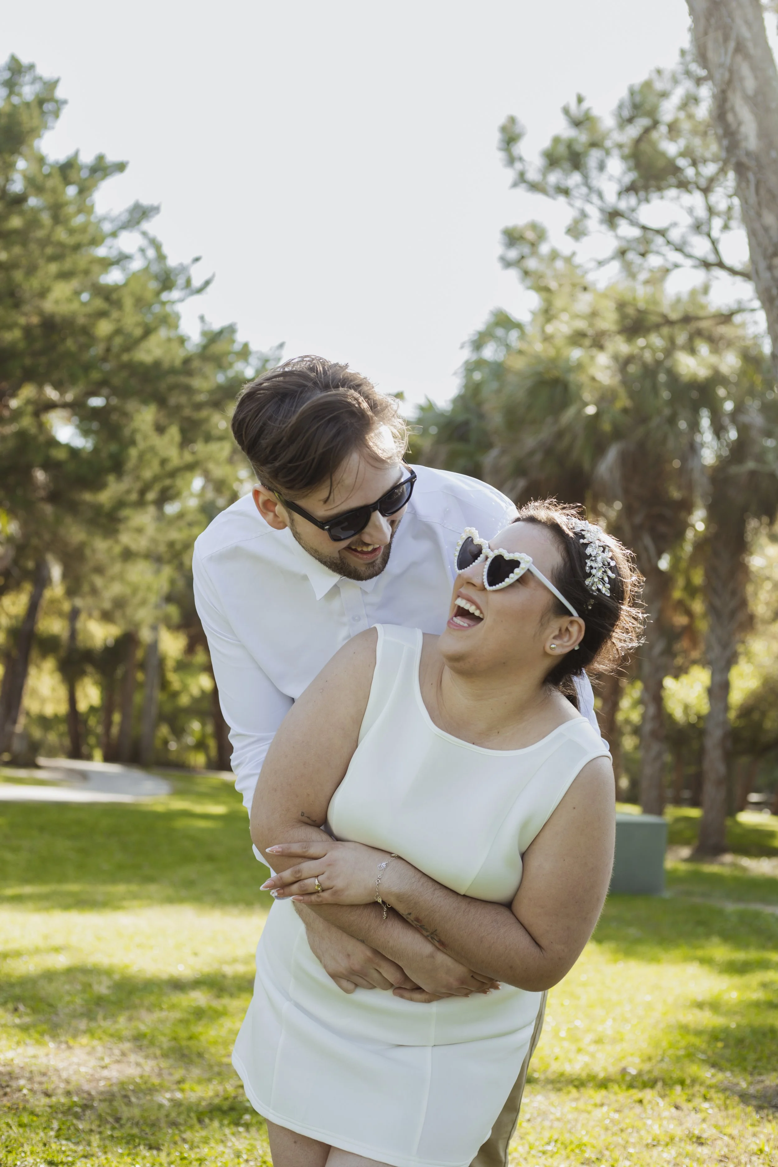 A man and a woman, both wearing sunglasses, sharing a joyful moment outdoors on a sunny day. The woman is wearing a white sleeveless dress and a floral headband, and the man is dressed in a white shirt. They are smiling and laughing, with trees and g