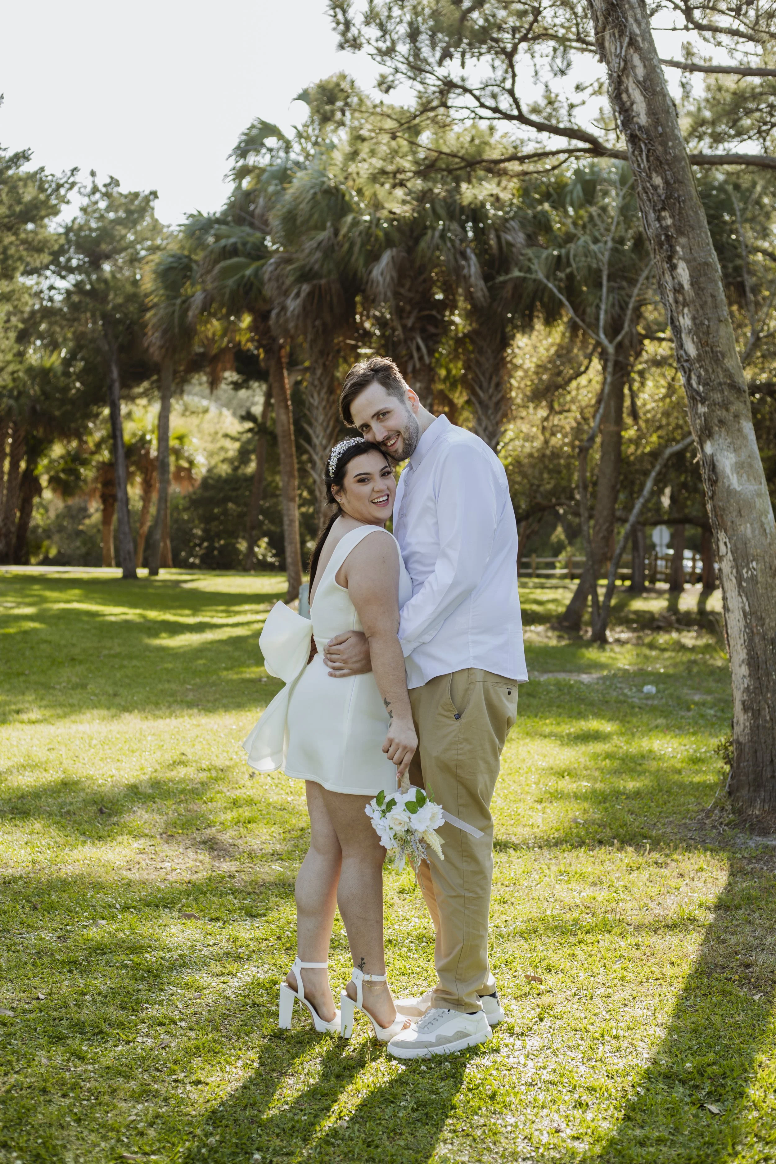 A happy couple standing outdoors in a park with lush green grass and tall trees, smiling and embracing each other, on a sunny day.