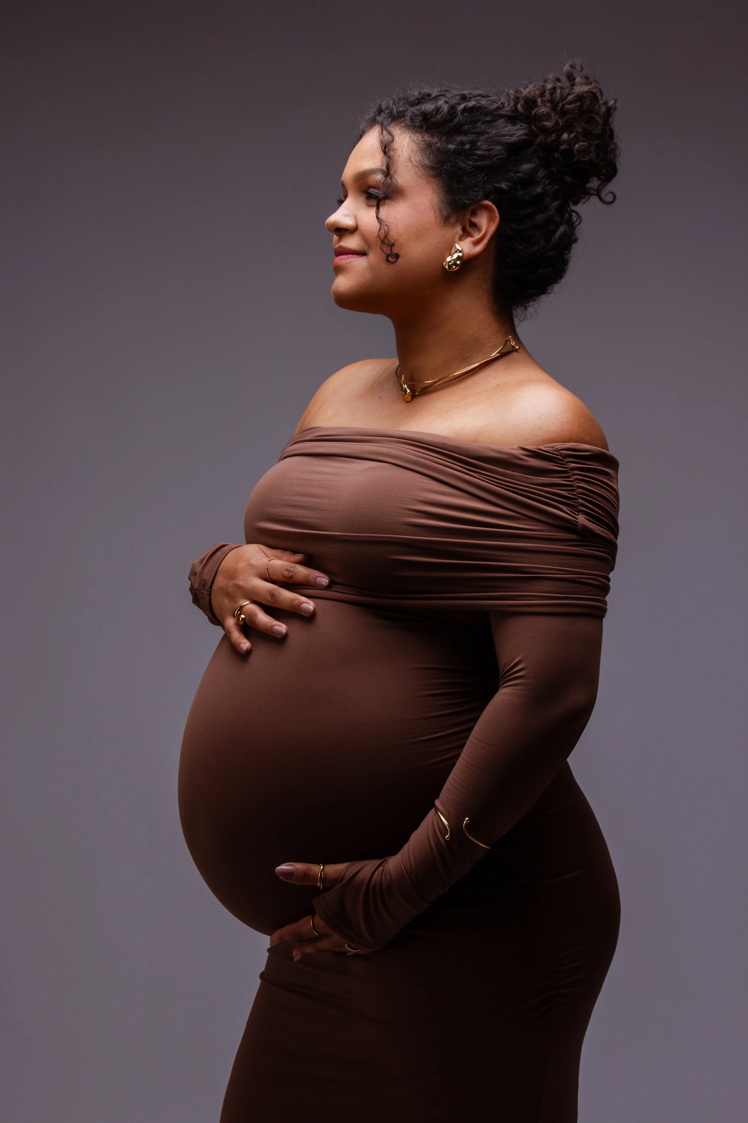 A pregnant woman with curly hair styled in a bun, wearing a brown off-shoulder dress and gold jewelry, standing sideways against a plain gray background.