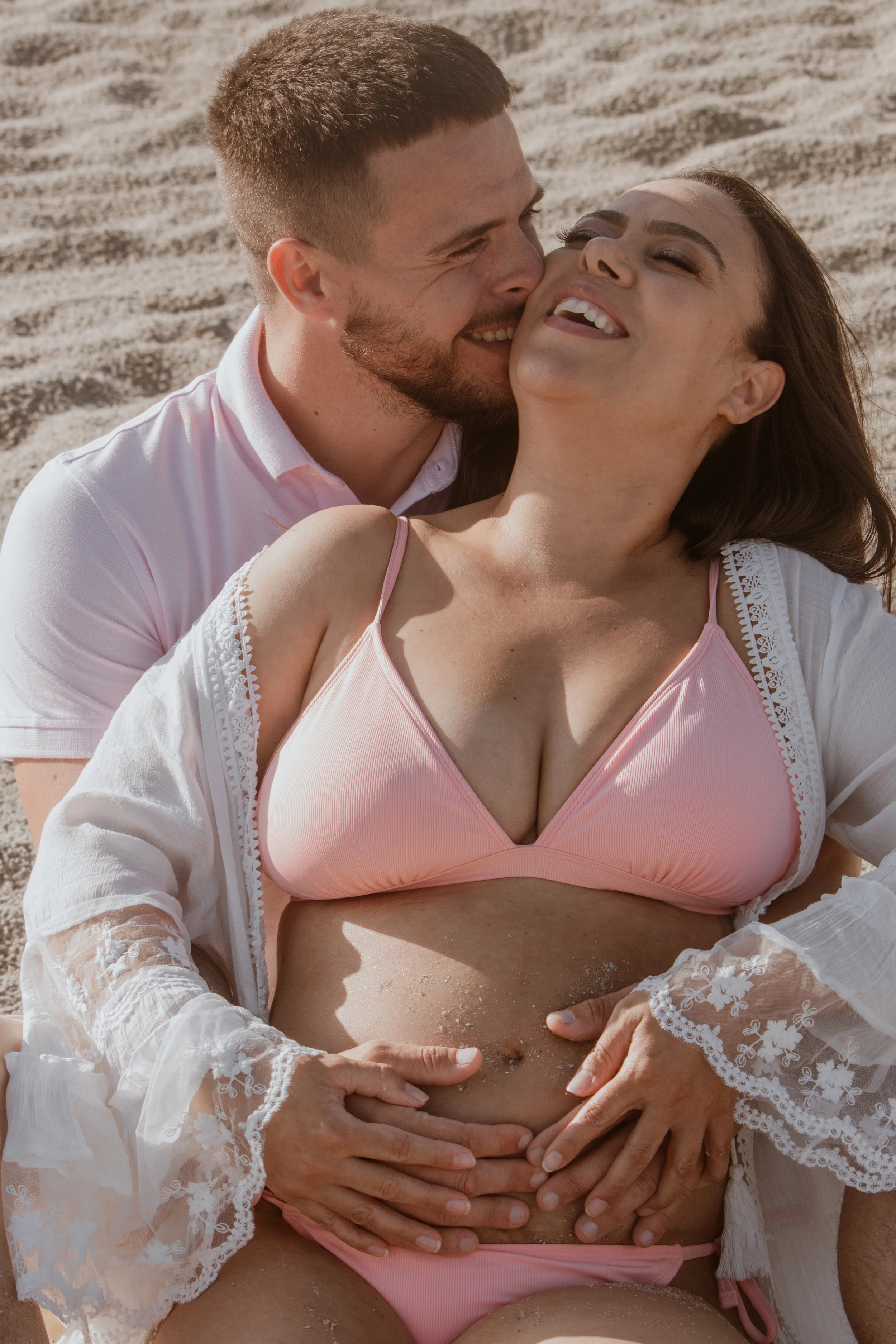 A smiling couple lying on the beach, the man embracing the woman from behind as she rests on the sand, both enjoying a sunny day.