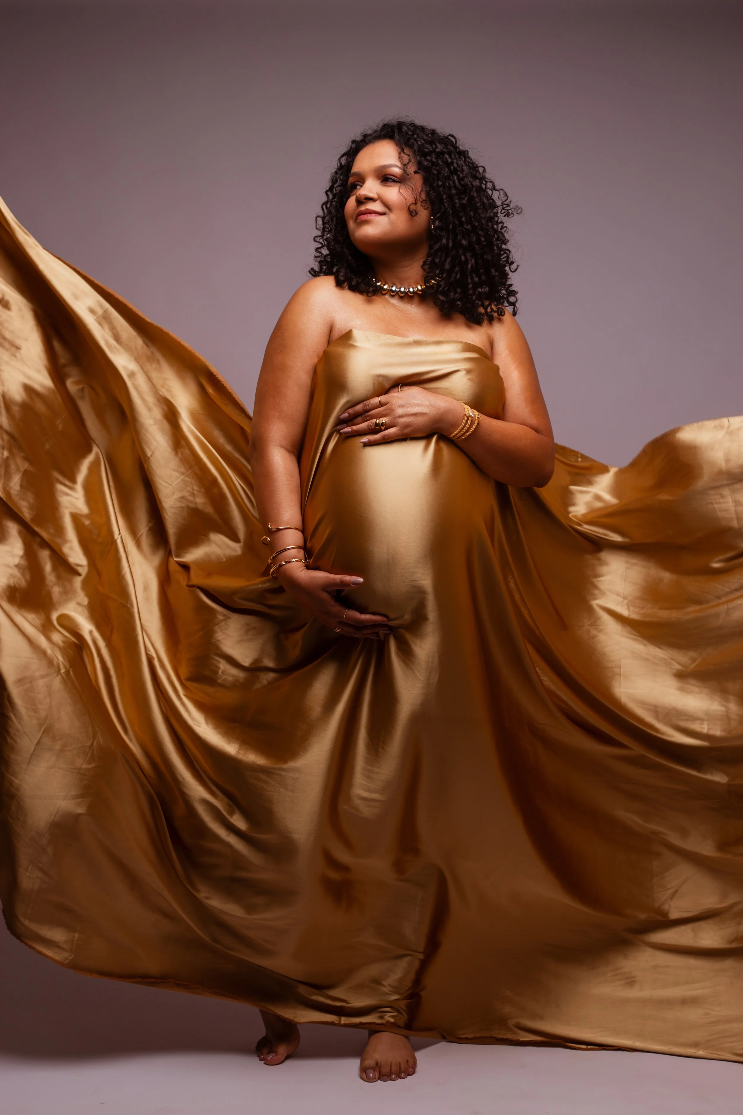 A pregnant woman with curly hair wearing a flowing gold satin dress, standing barefoot against a neutral background, with one hand on her belly and the other hand resting on her chest, smiling softly.