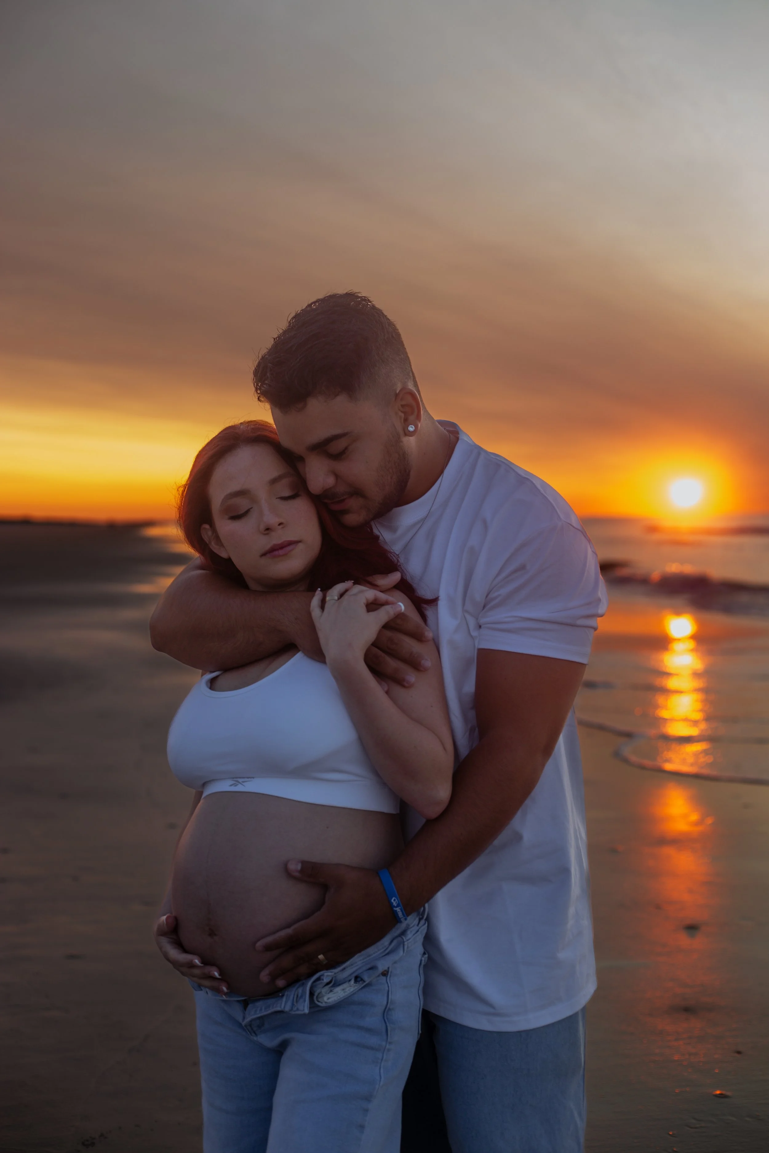 A pregnant woman and a man embracing on a beach at sunset