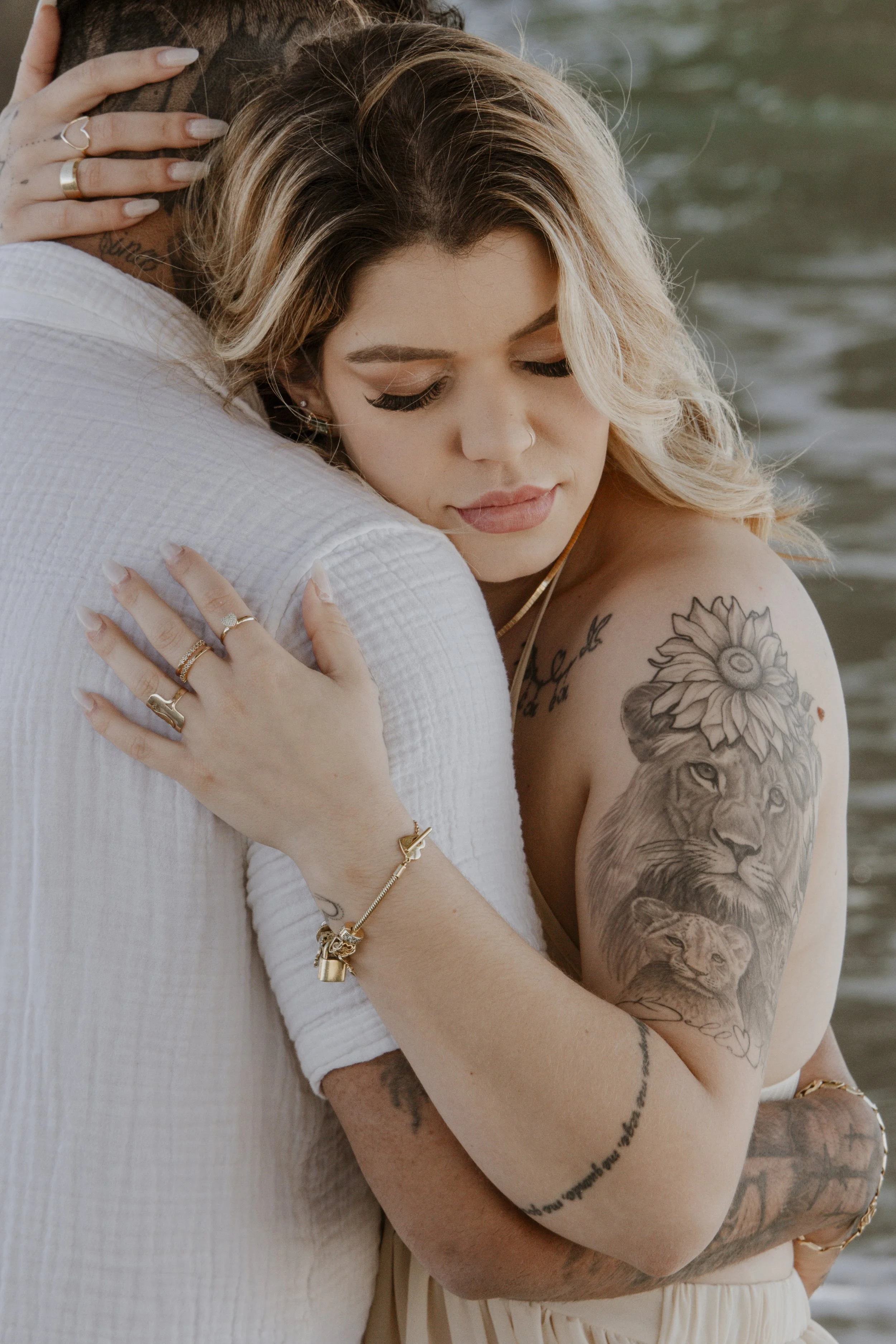 A woman with tattoos, blonde hair, and makeup embracing a man near the water, both appearing to be happy.