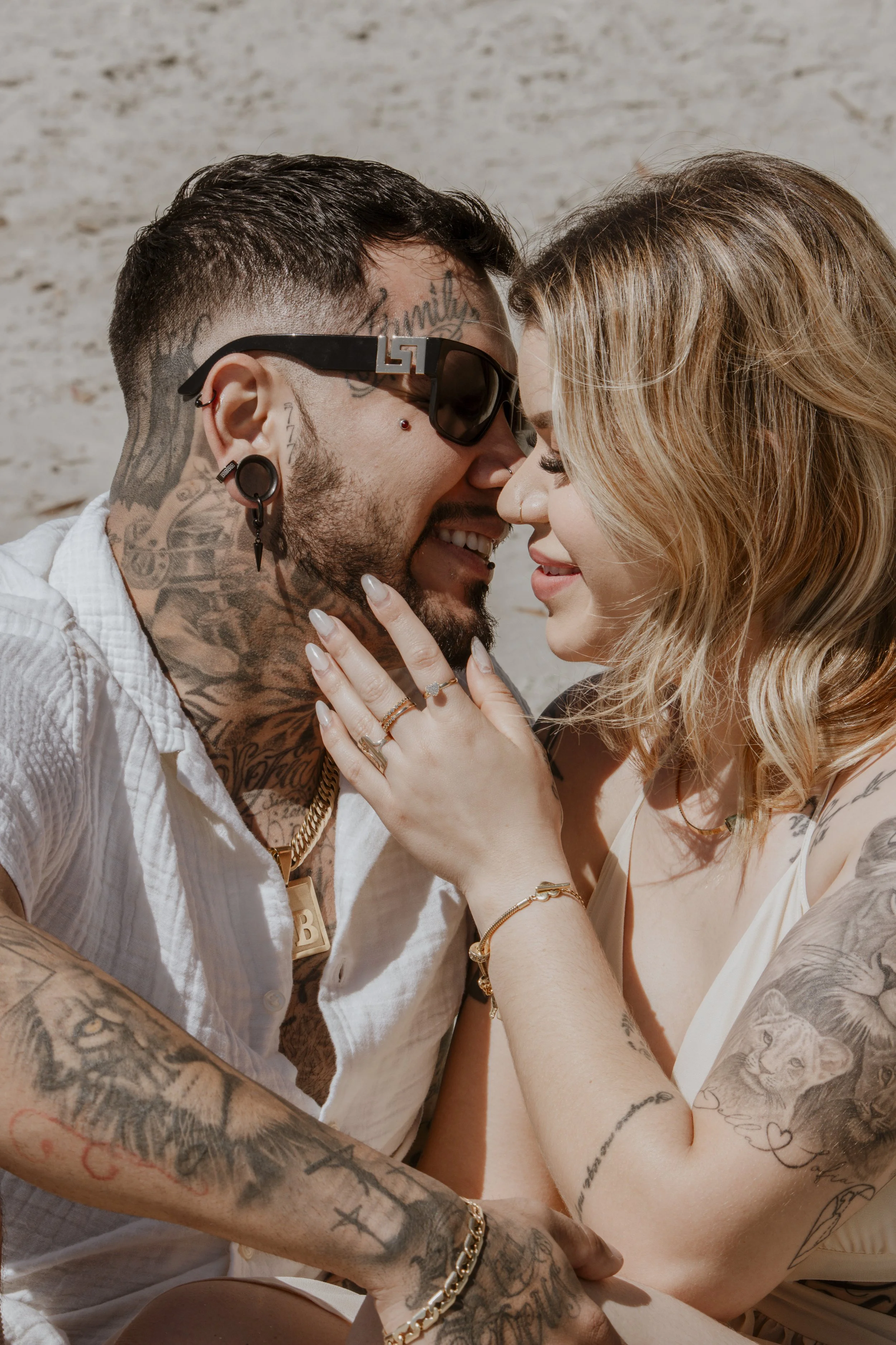 A close-up of a smiling couple touching foreheads and noses, with tattoos and jewelry, on a beach.