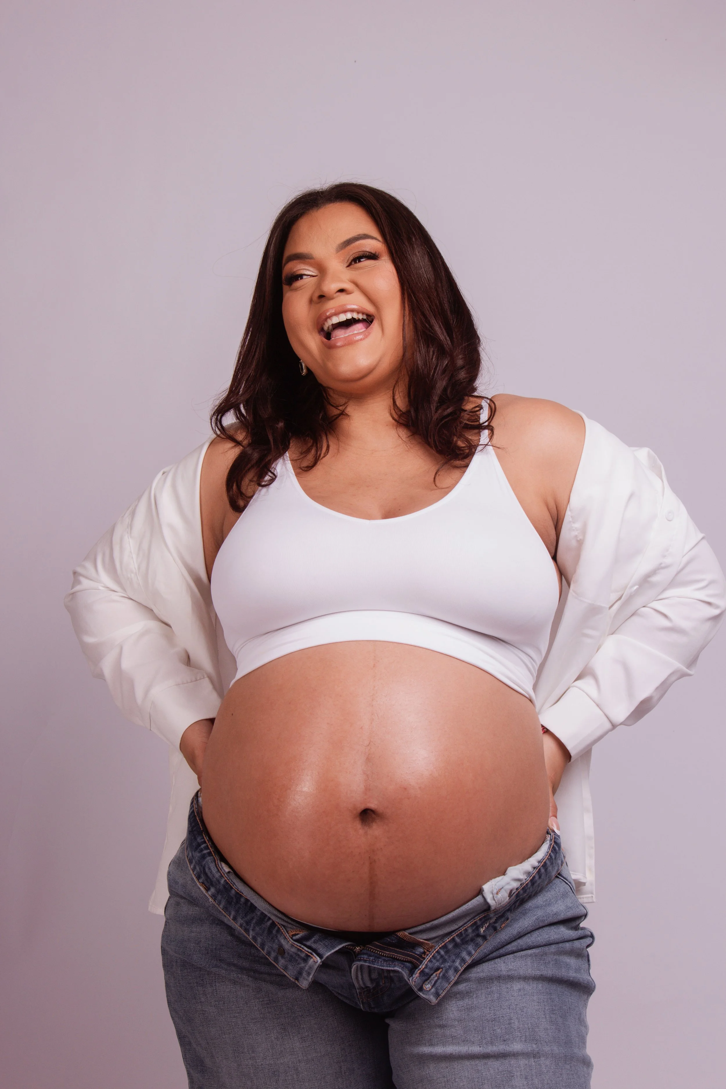 A pregnant woman with a big belly, wearing a white sports bra and gray jeans, smiling and laughing.