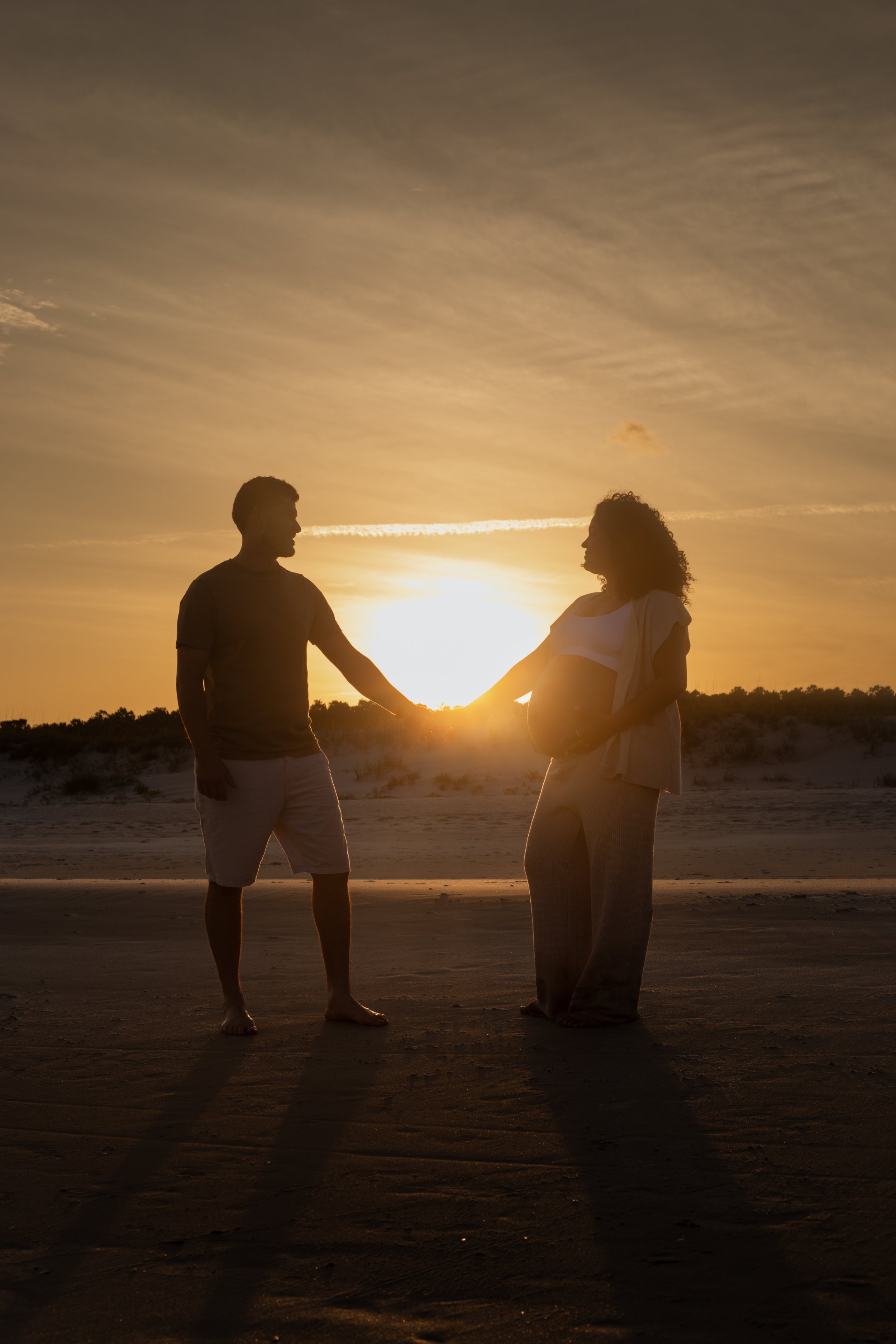 Silhouette of a pregnant woman and a man holding hands on the beach at sunset, with the sun behind them and their shadows on the sand.
