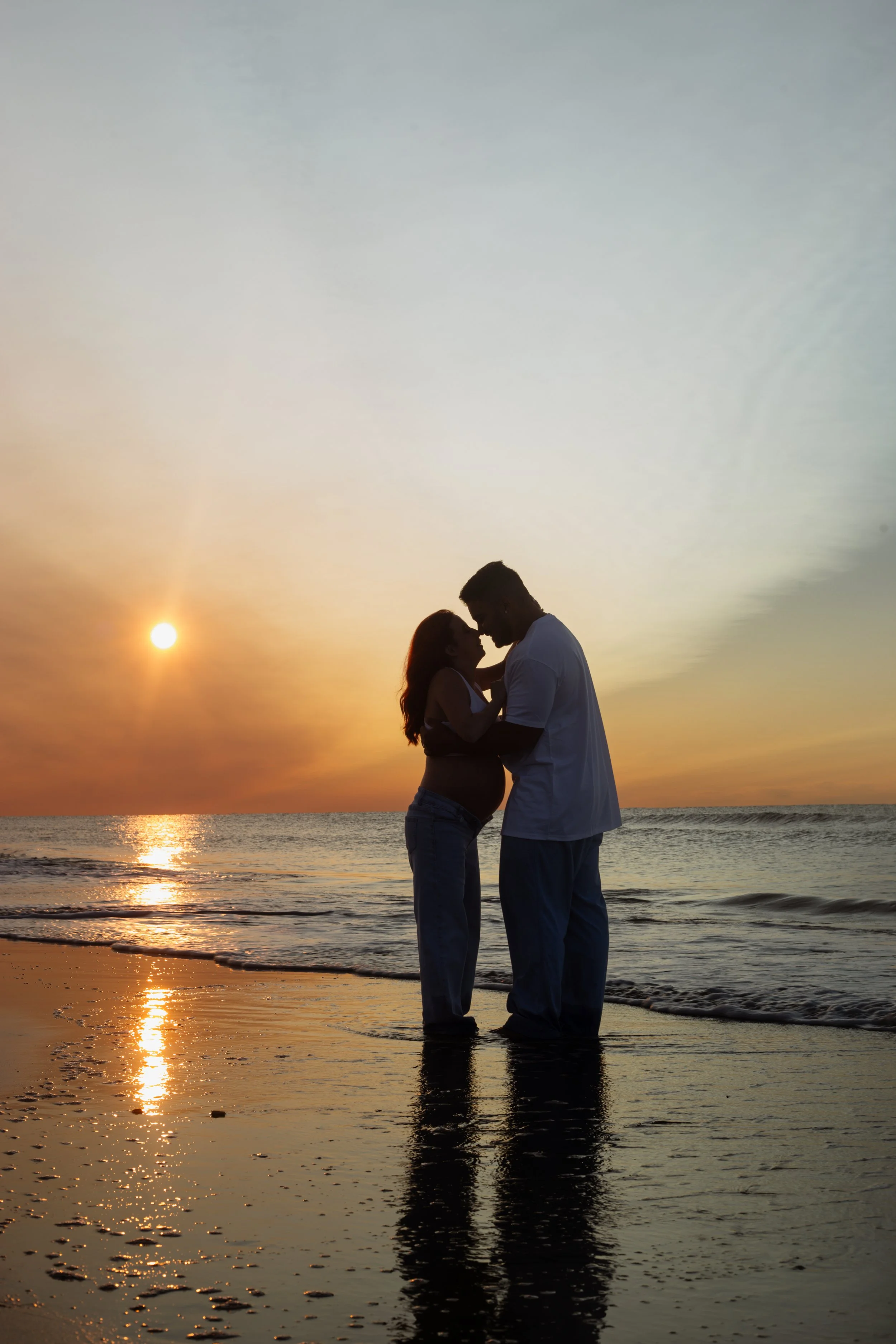Silhouette of a couple embracing and touching foreheads at sunset on the beach, with the ocean and colorful sky in the background.