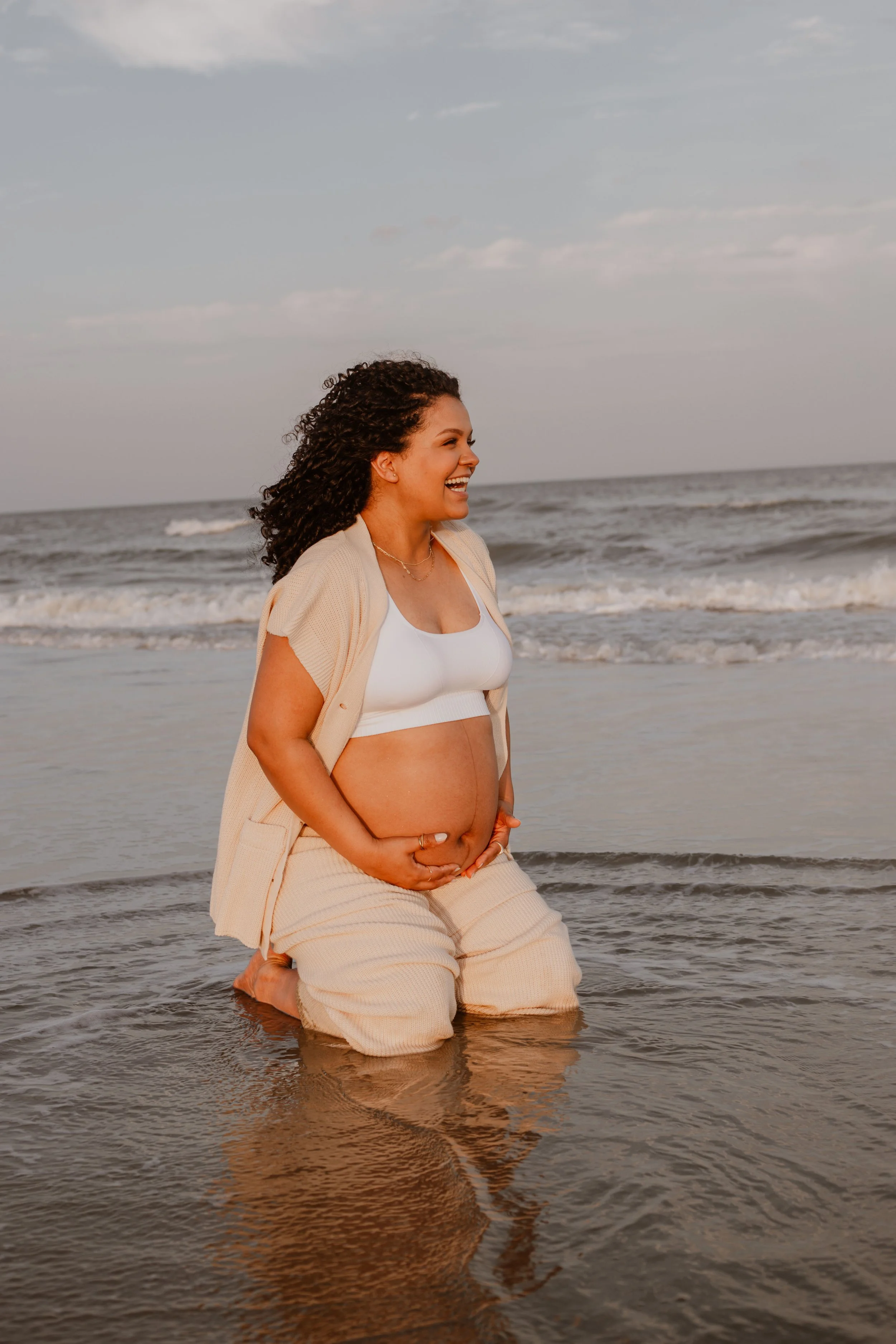 A pregnant woman kneels in shallow ocean water at the beach, smiling and holding her belly, during sunset.