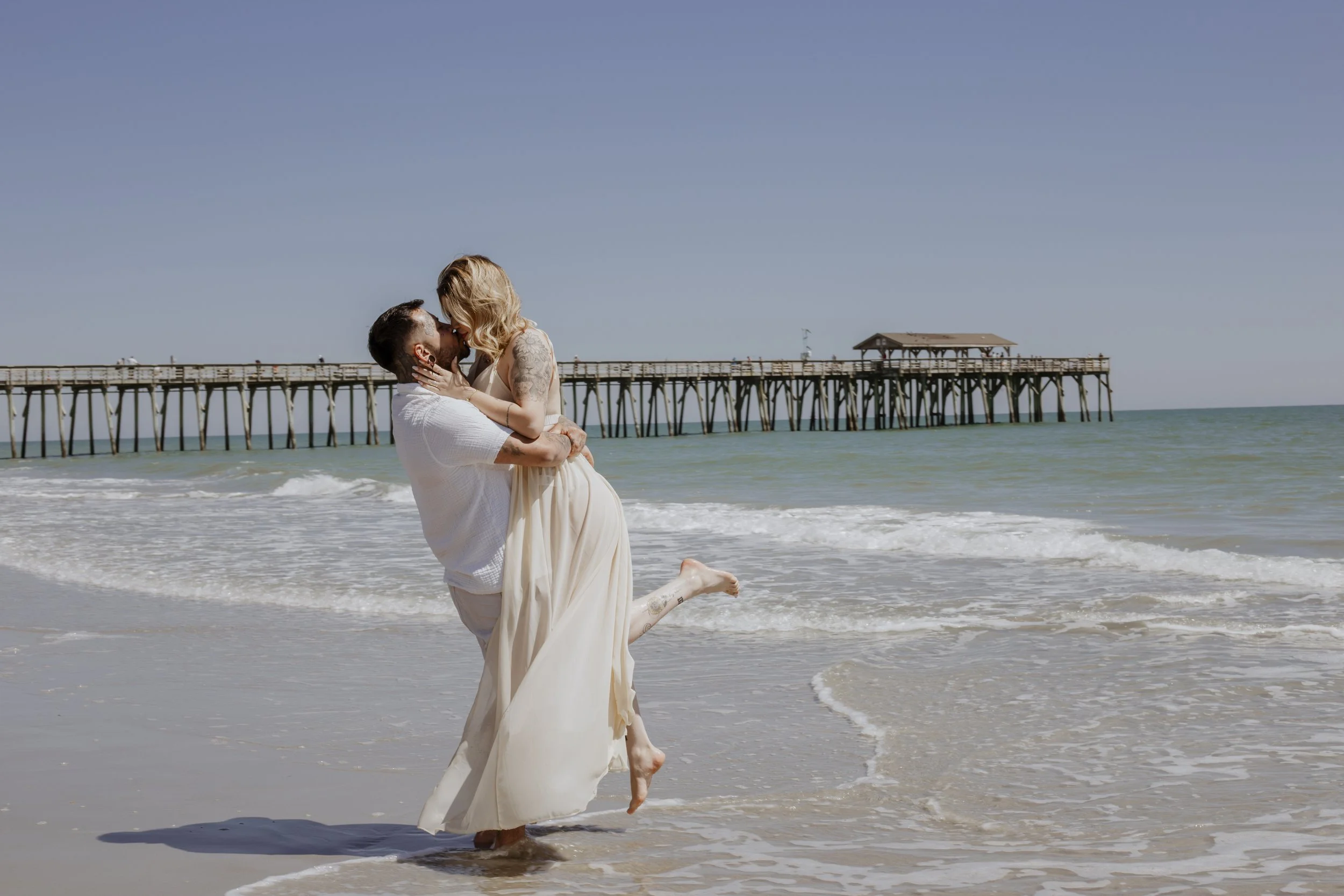 A couple kissing on the beach with a pier in the background.