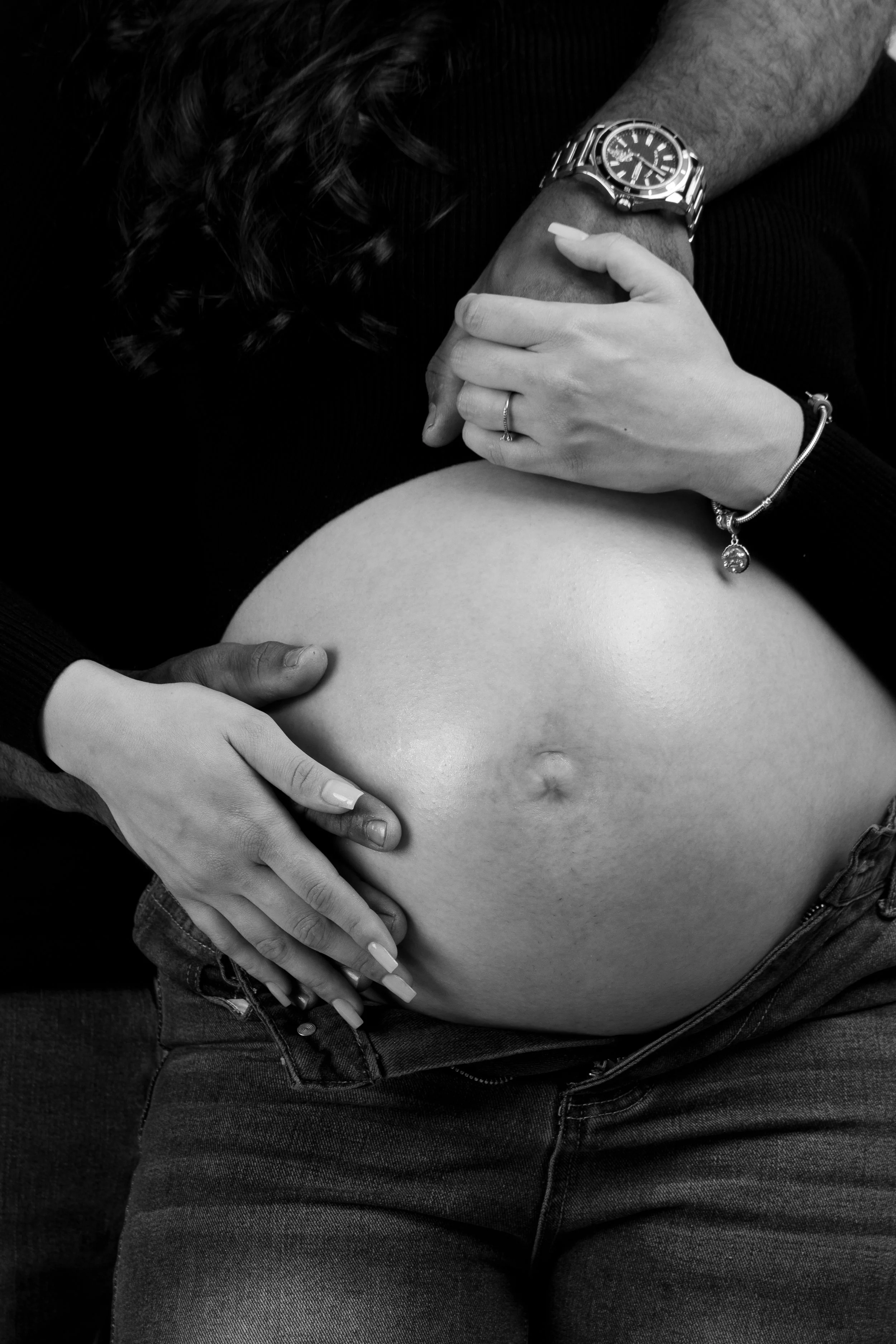 Black and white photo of a pregnant woman’s belly, with hands and a man’s hand resting on it. The woman's nails are manicured, and they wear a bracelet and a ring. The man is wearing a watch.