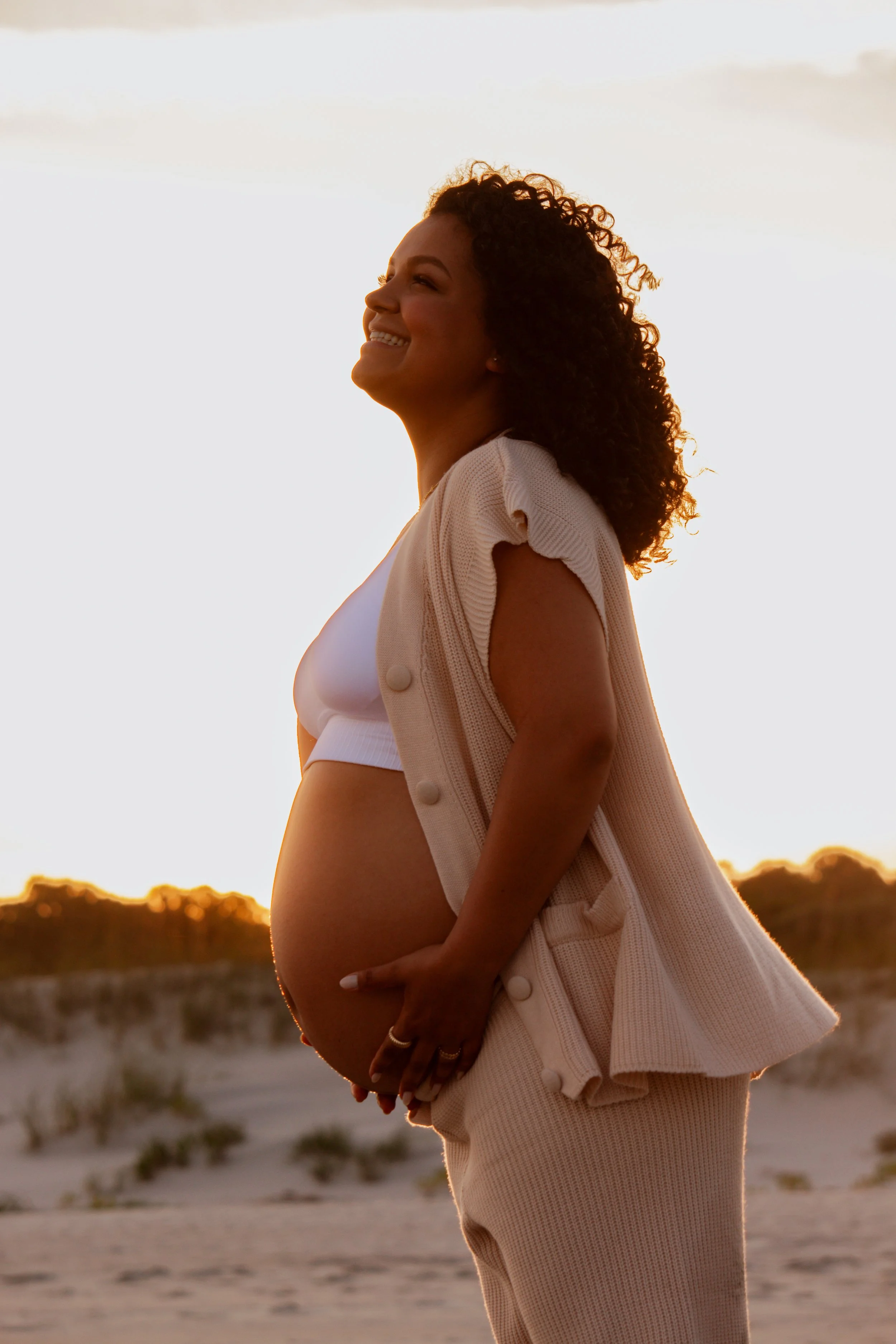 Pregnant woman smiling with hands on belly, standing on a beach at sunset.