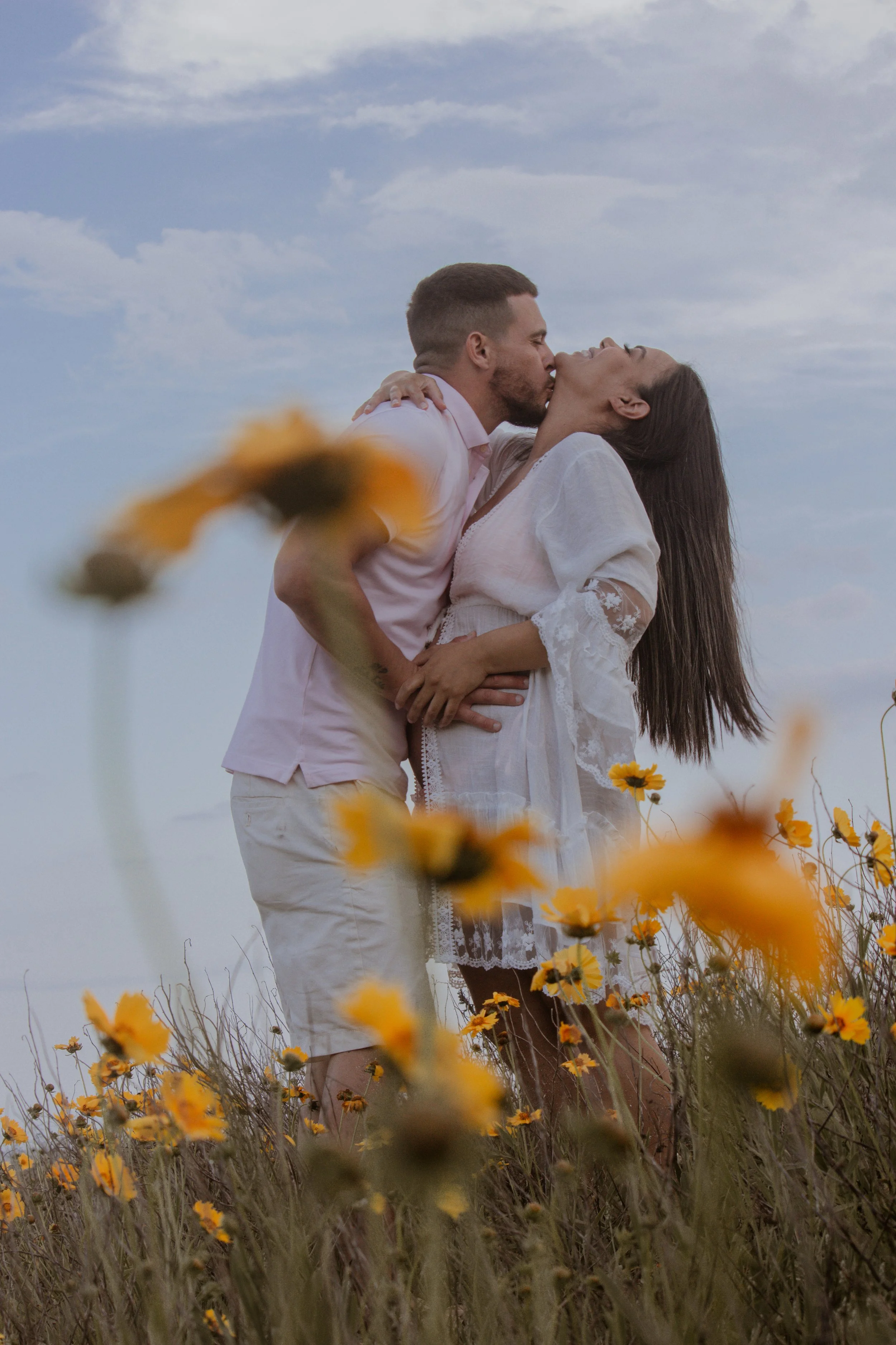 A couple is kissing in a yellow flower meadow under a partly cloudy sky.