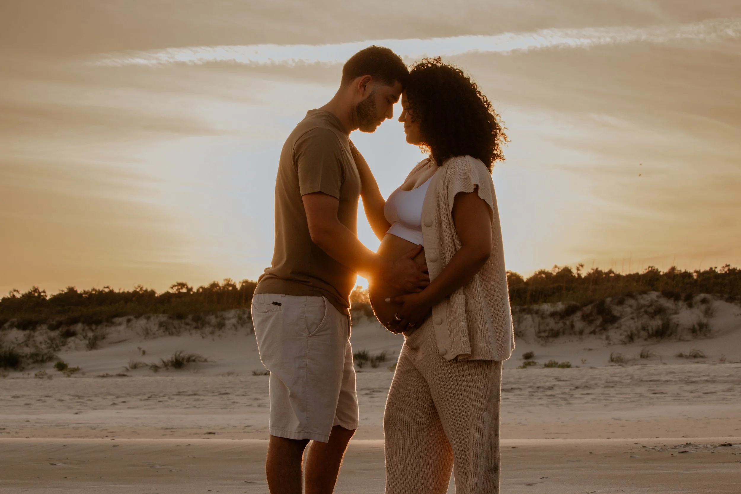 A pregnant woman and her partner standing on the beach at sunset, with their foreheads touching, and the partner touching her belly.