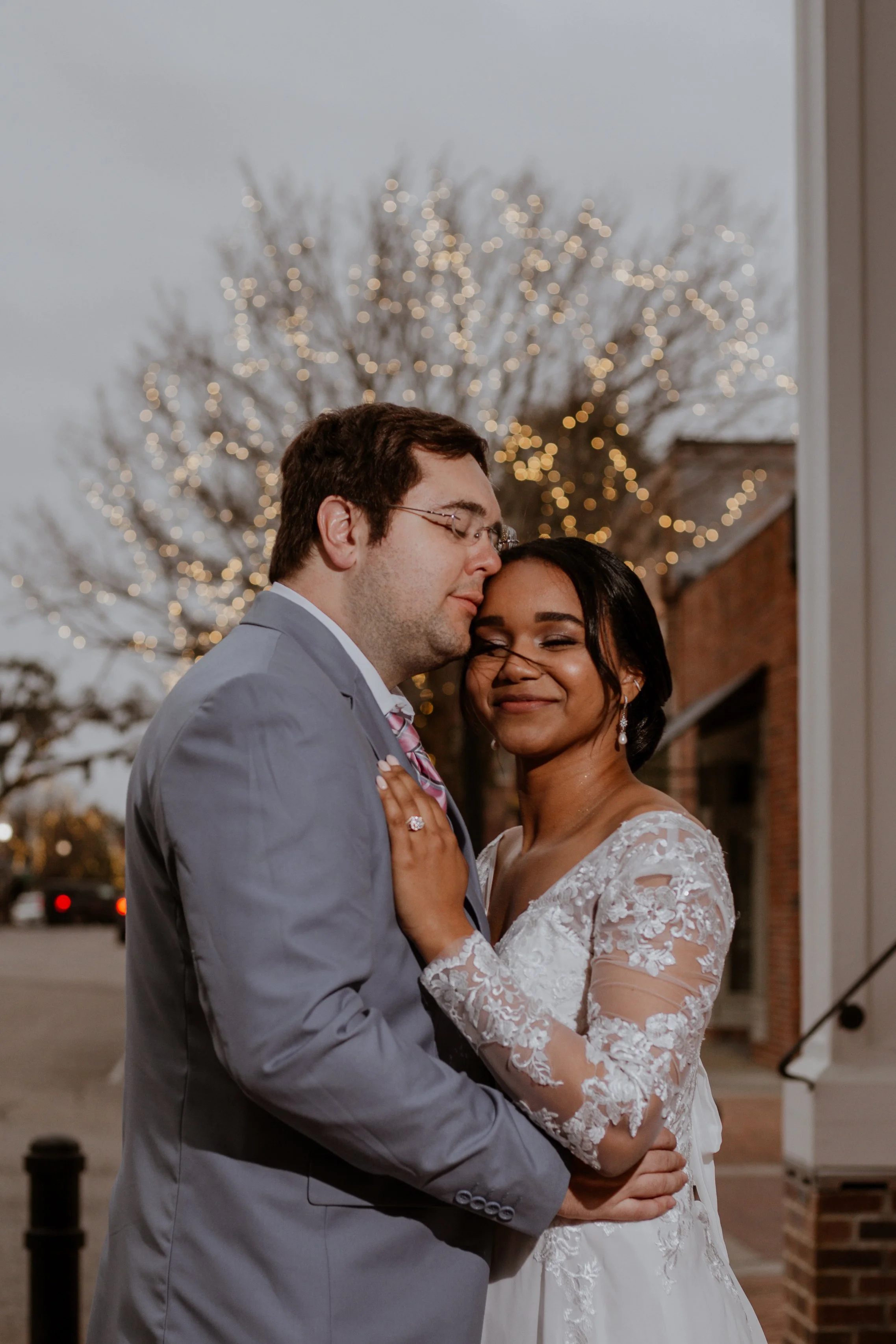 A newlywed couple embraces outside during dusk, with a tree decorated with string lights in the background.