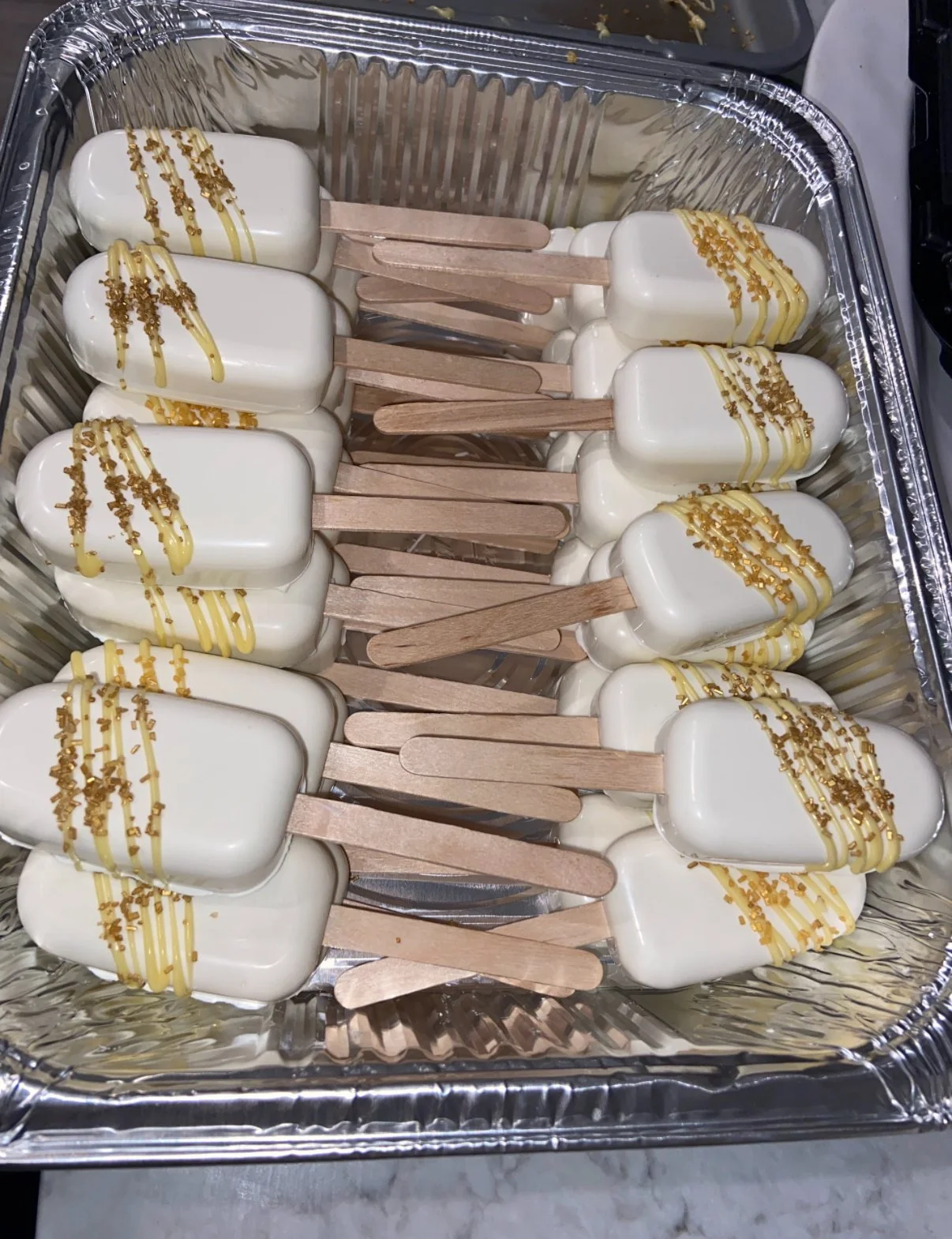 Several white popsicles with wooden sticks, decorated with yellow drizzle and gold sprinkles, arranged in a tray.