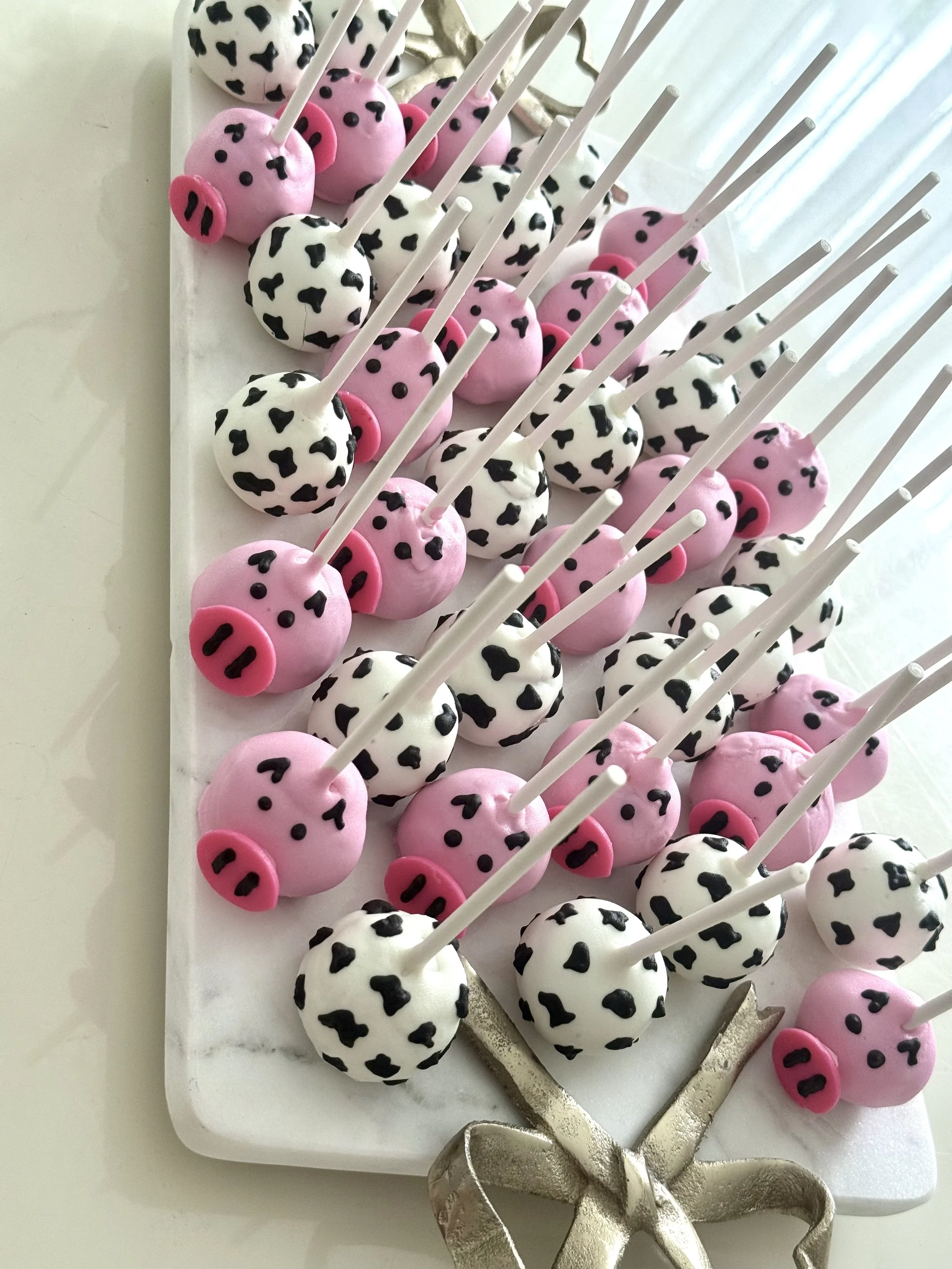Cake pops decorated as cow and pig faces with black and white spots and pink snouts, arranged on a white tray with a silver ribbon bow at the corner.