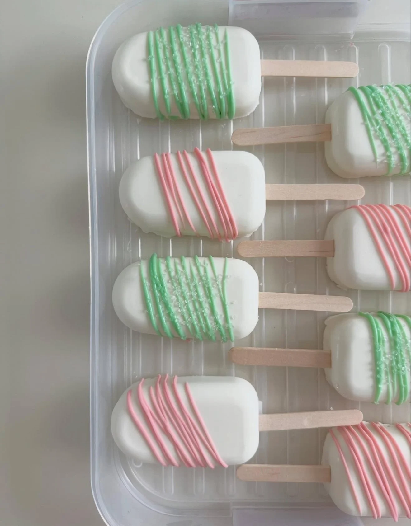 Cream-colored popsicles with pink and green drizzle and sprinkles, arranged on a clear tray.