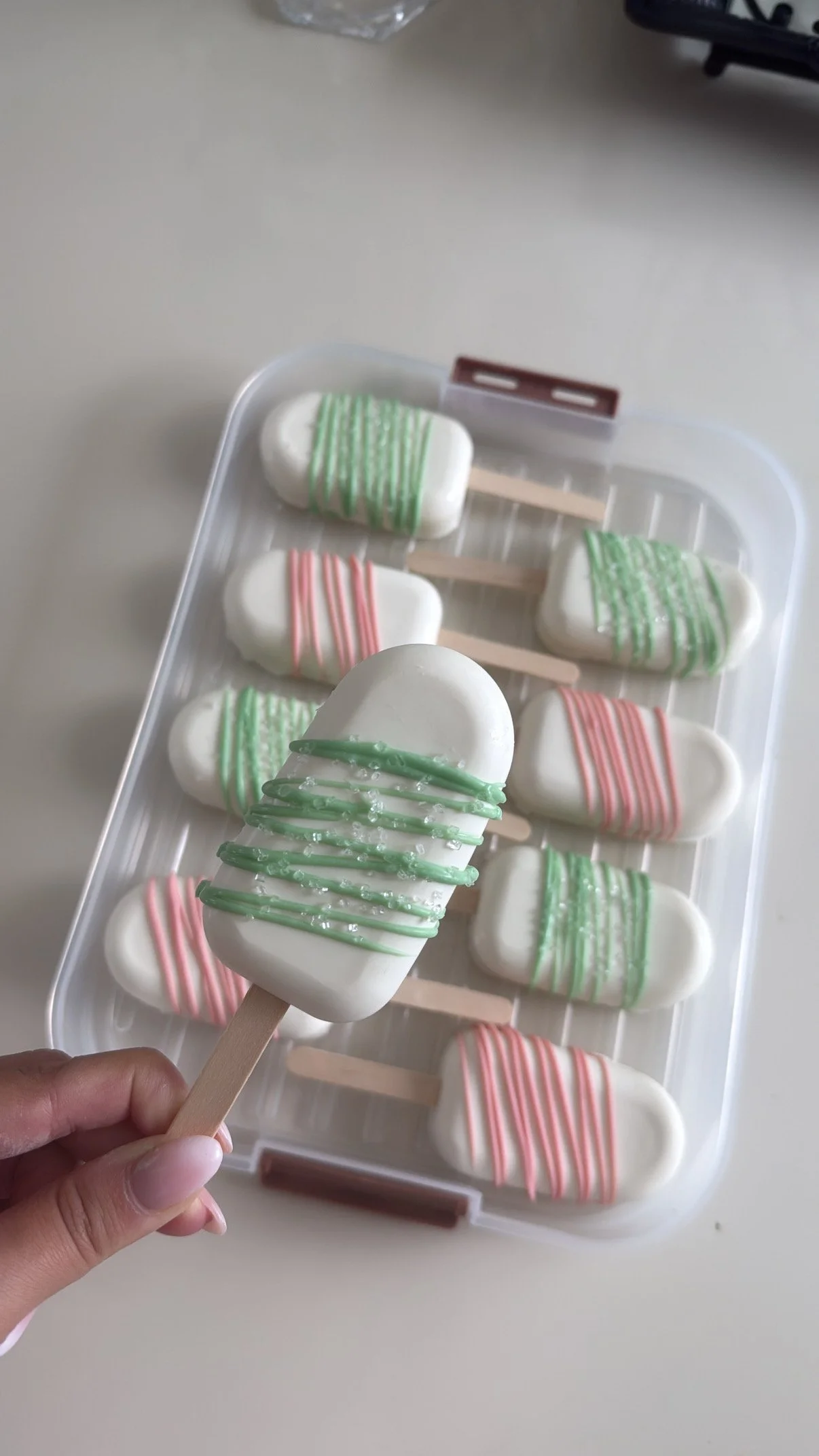 Celebration ice pops with white coating, green and pink drizzle, and sparkles in a plastic container.