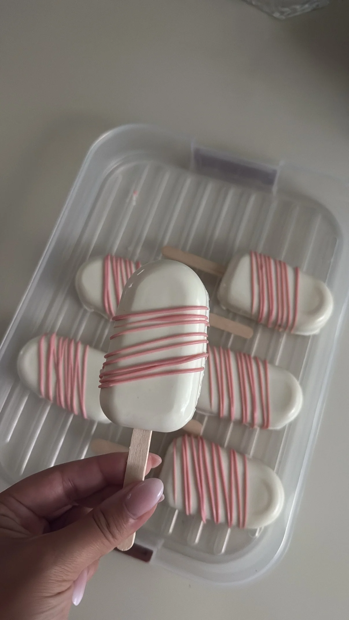 Hand holding a white, rectangular ice cream bar with pink drizzle on a wooden stick, with six similar ice cream bars in the background on a plastic tray.