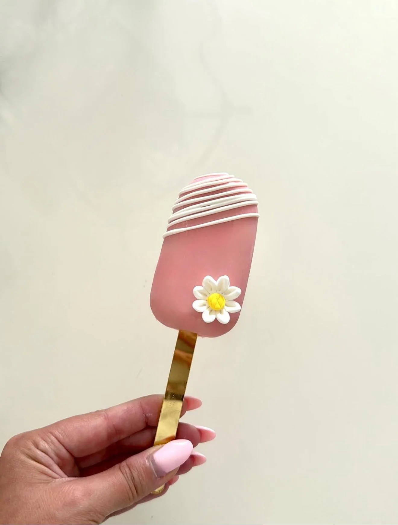 Pink ice cream bar with white drizzle and a small white flower decoration on top, held by a hand with manicured nails, against a plain light background.