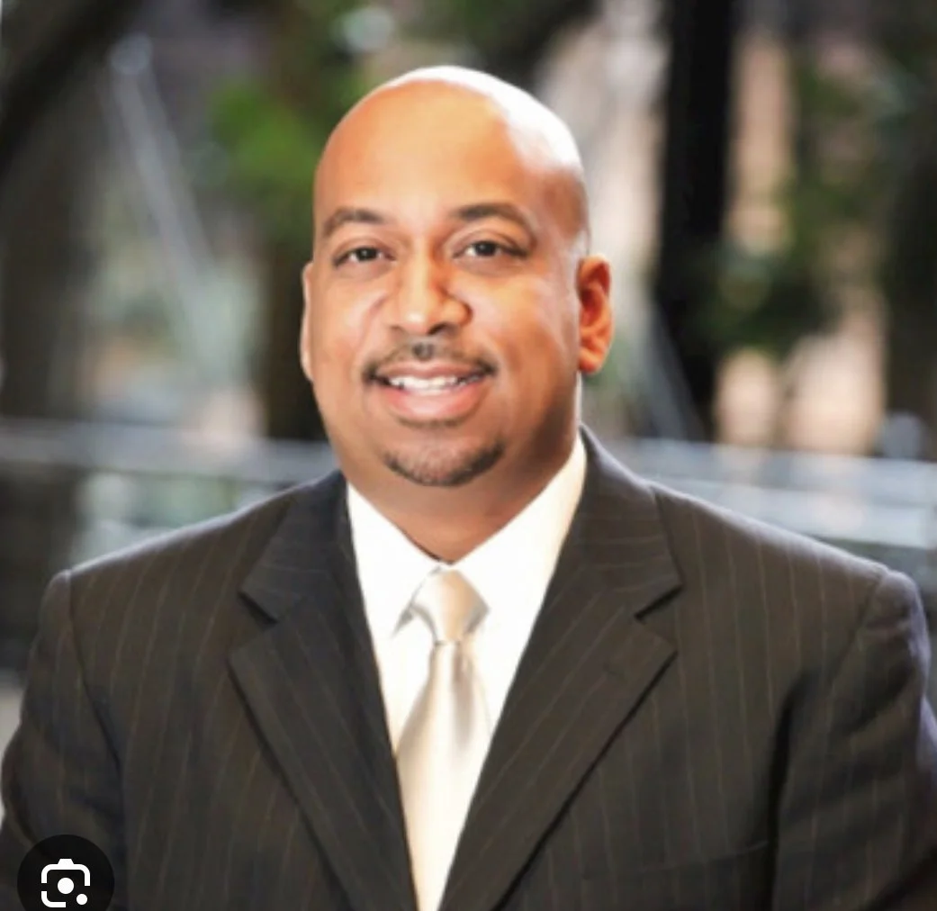 A man in a dark pinstripe suit, white shirt, and beige tie, smiling outdoors with blurred trees in the background.