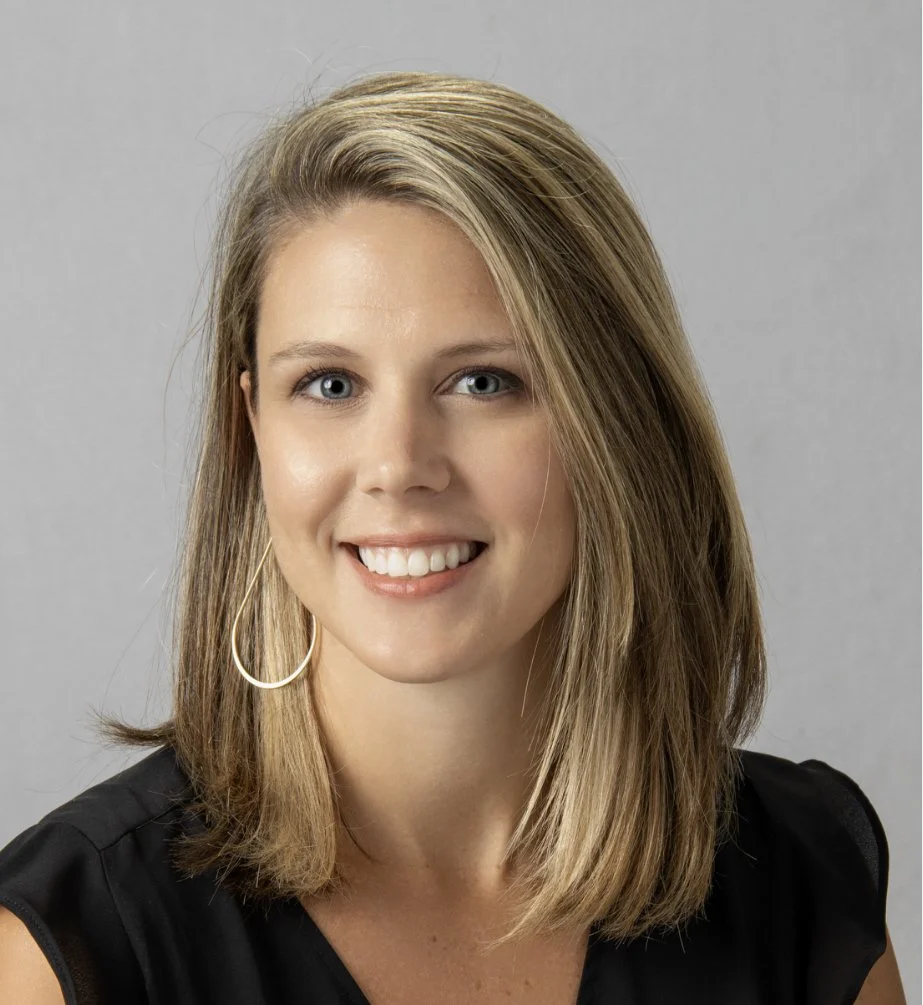 A woman smiling with shoulder-length blonde hair, wearing a black top and large hoop earrings, against a plain gray background.