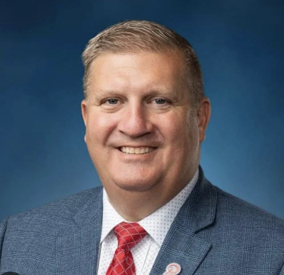 Portrait of a smiling man with blonde hair wearing a gray plaid suit, white shirt, and red patterned tie, against a blue background.