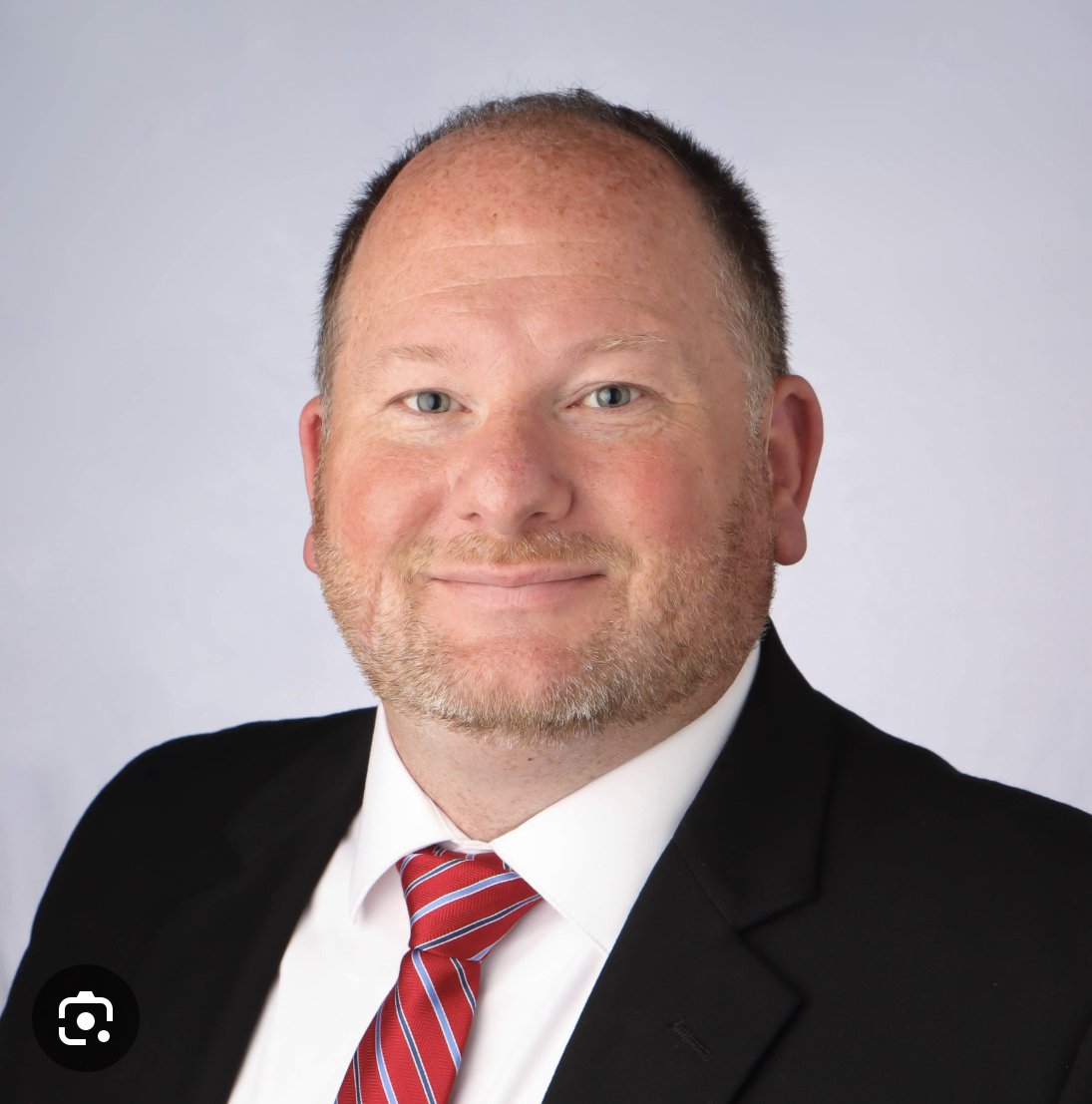 A middle-aged man with short, light-colored hair and a beard wearing a black suit, white shirt, and a red striped tie, smiling against a plain gray background.