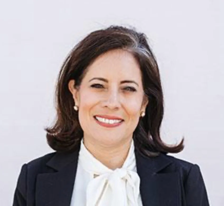 Portrait of a woman in a business suit with a white blouse and pearl earrings, smiling against a plain light background.