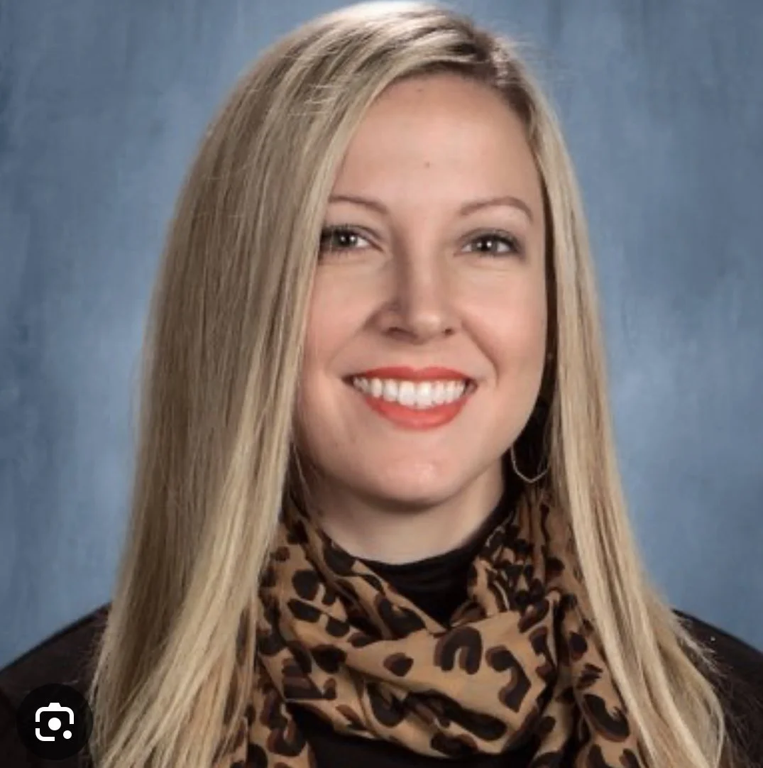 A woman with long blonde hair smiling, wearing a black top and a leopard-print scarf, posed against a blue background.