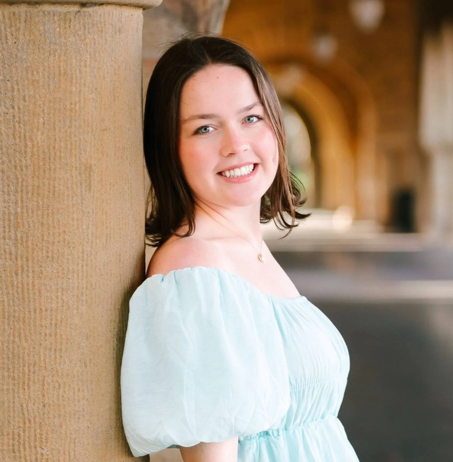 A young woman with shoulder-length dark hair, wearing a light blue off-the-shoulder dress, smiling and leaning against a stone pillar in an outdoor setting with arches in the background.
