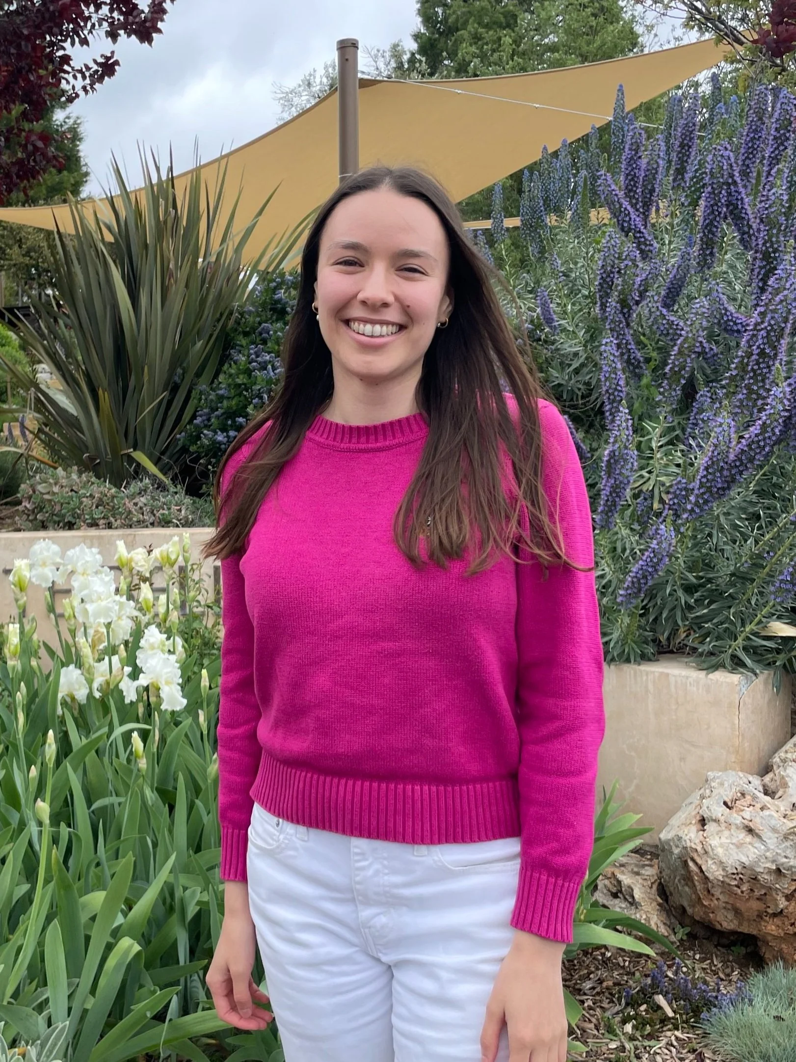 A young woman with long brown hair smiling outdoors, wearing a pink sweater and white pants, standing in front of purple and white flowers with a yellow shade sail overhead.