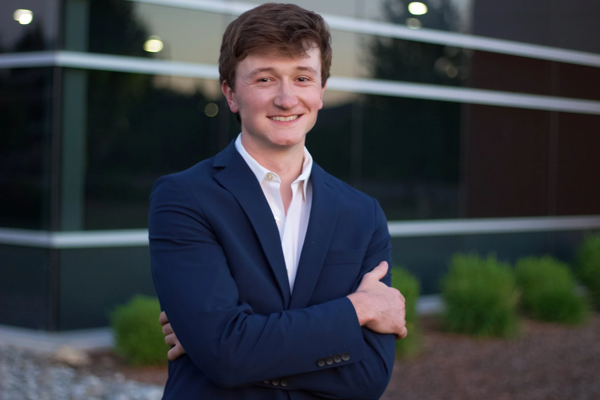 A young man in a navy blue suit with a white shirt, smiling with arms crossed, standing outdoors in front of a modern glass building with greenery.