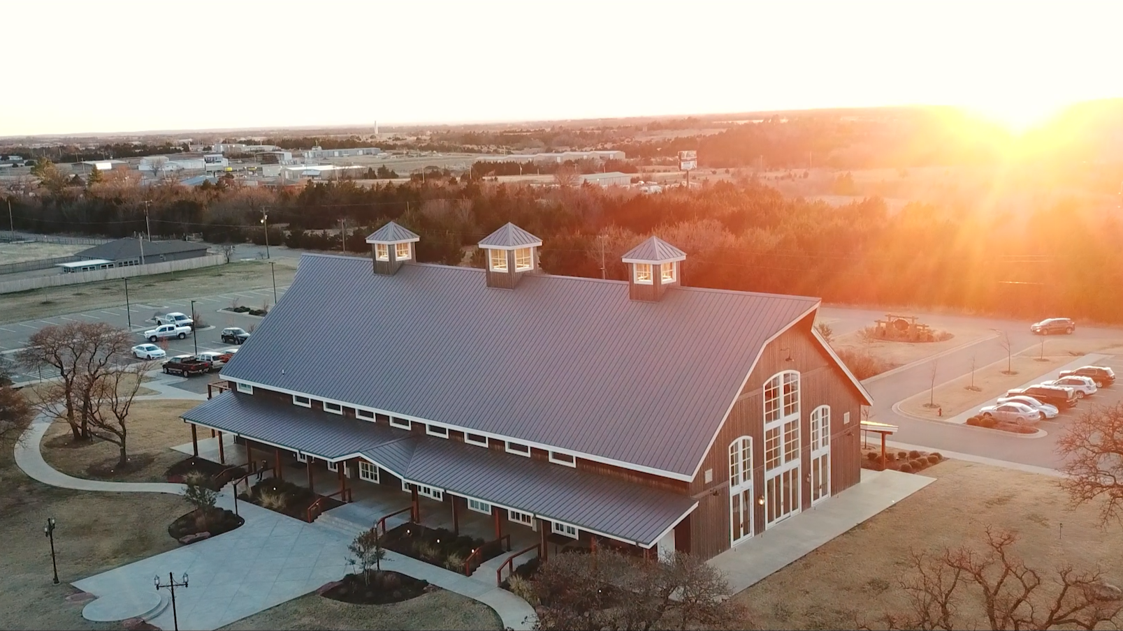 A large barn-style building with a metal roof and three small turrets on top, illuminated by the setting sun, surrounded by a parking lot and landscaped area with trees and winding walkways.