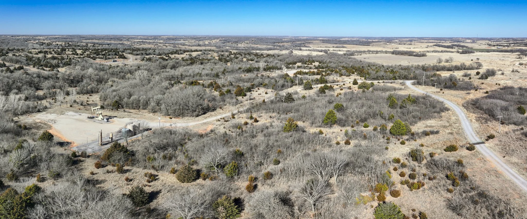 Aerial view of a rural landscape with open fields, sparse trees, a winding dirt road, and a small oil pumpjack.
