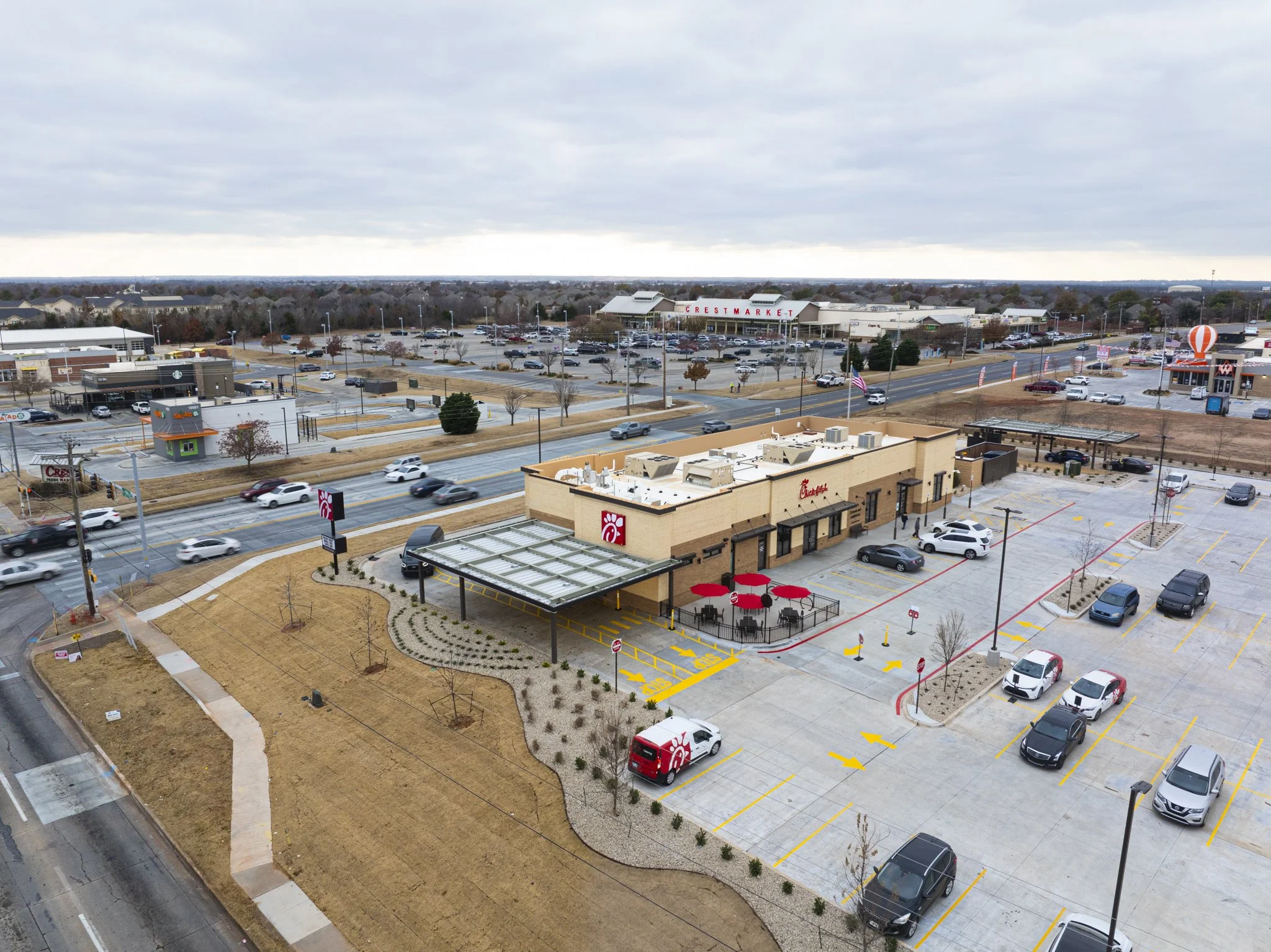 Aerial view of a Chick-fil-A restaurant with outdoor seating, parking lot, and surrounding shopping area on an overcast day.