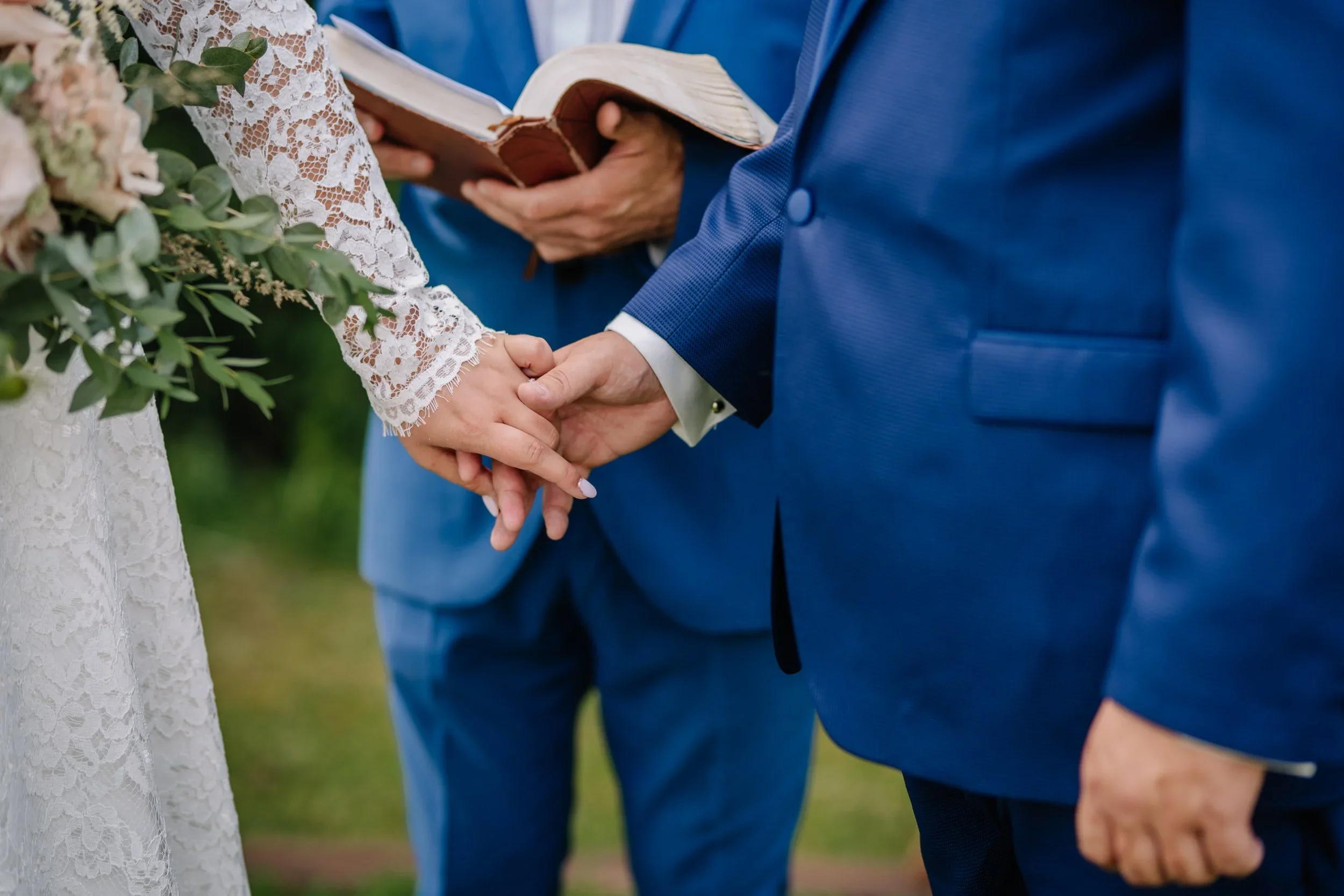A couple holding hands during a wedding ceremony, with a person in a blue suit reading from a book.