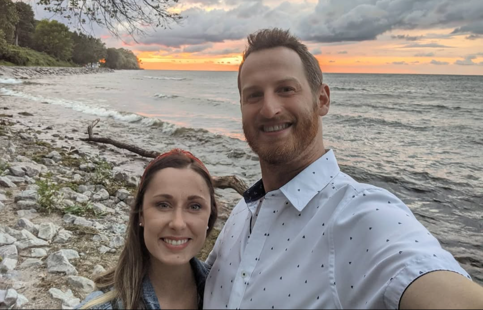 A smiling couple taking a selfie on a rocky beach during sunset, with the ocean and colorful sky in the background.