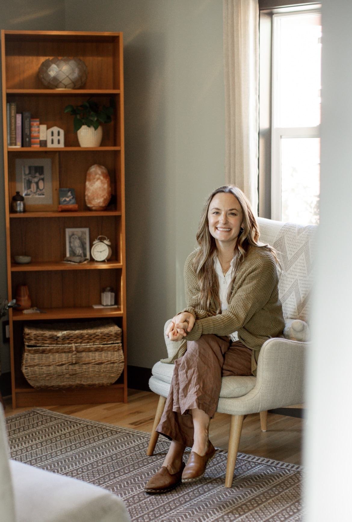 A woman with long light brown hair sitting on a beige armchair in a living room, smiling at the camera. She is wearing a white shirt, brown pants, a tan cardigan, and brown shoes. The room features a wooden bookshelf with decorative items, photos, and books, next to a window with cream curtains, and a patterned rug on the floor.
