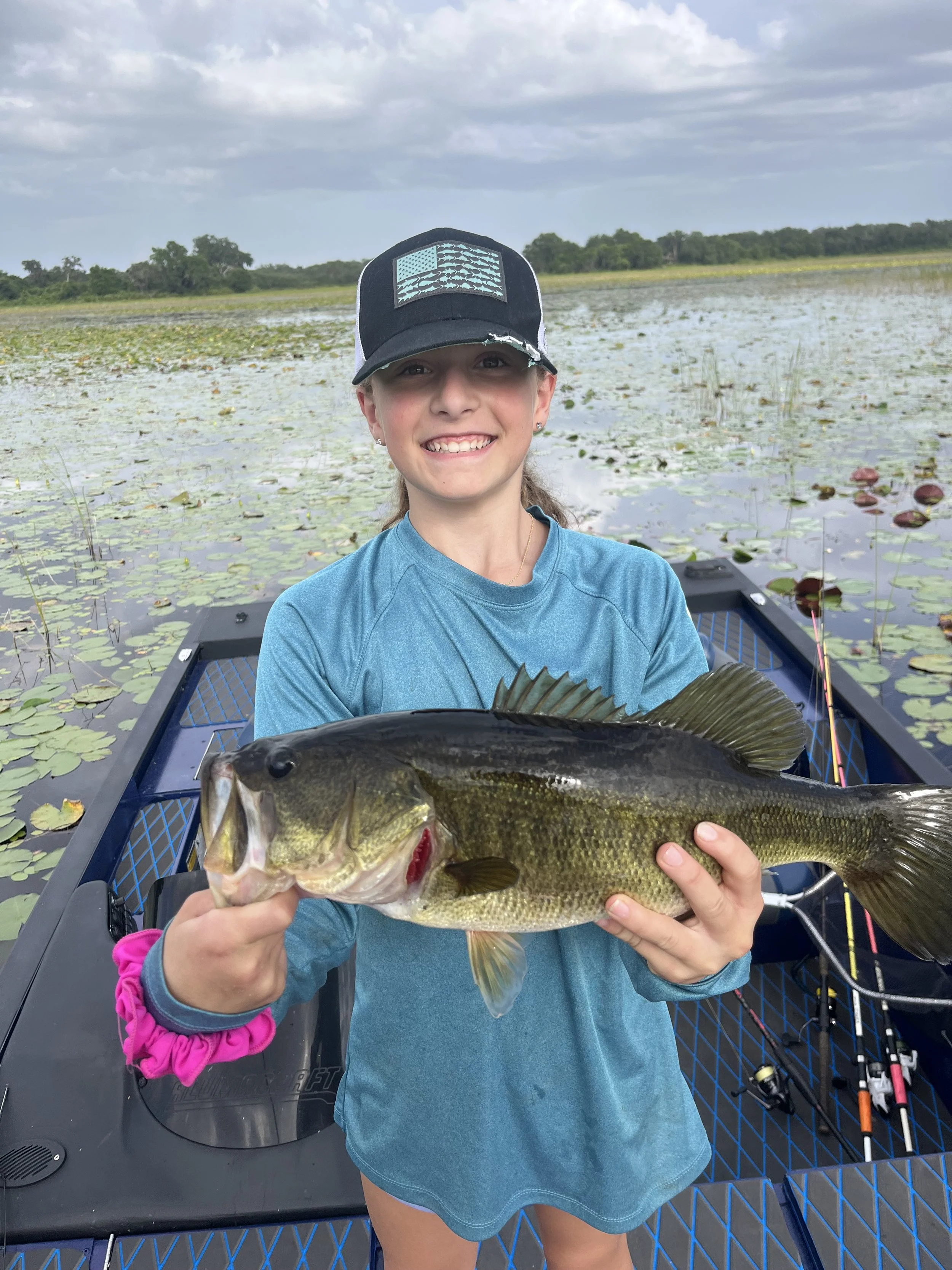 4lb Bass Young girl in a blue long-sleeve shirt holding a large fish she caught while on a boat in a marshy area with lily pads and trees in the background, smiling at the camera.