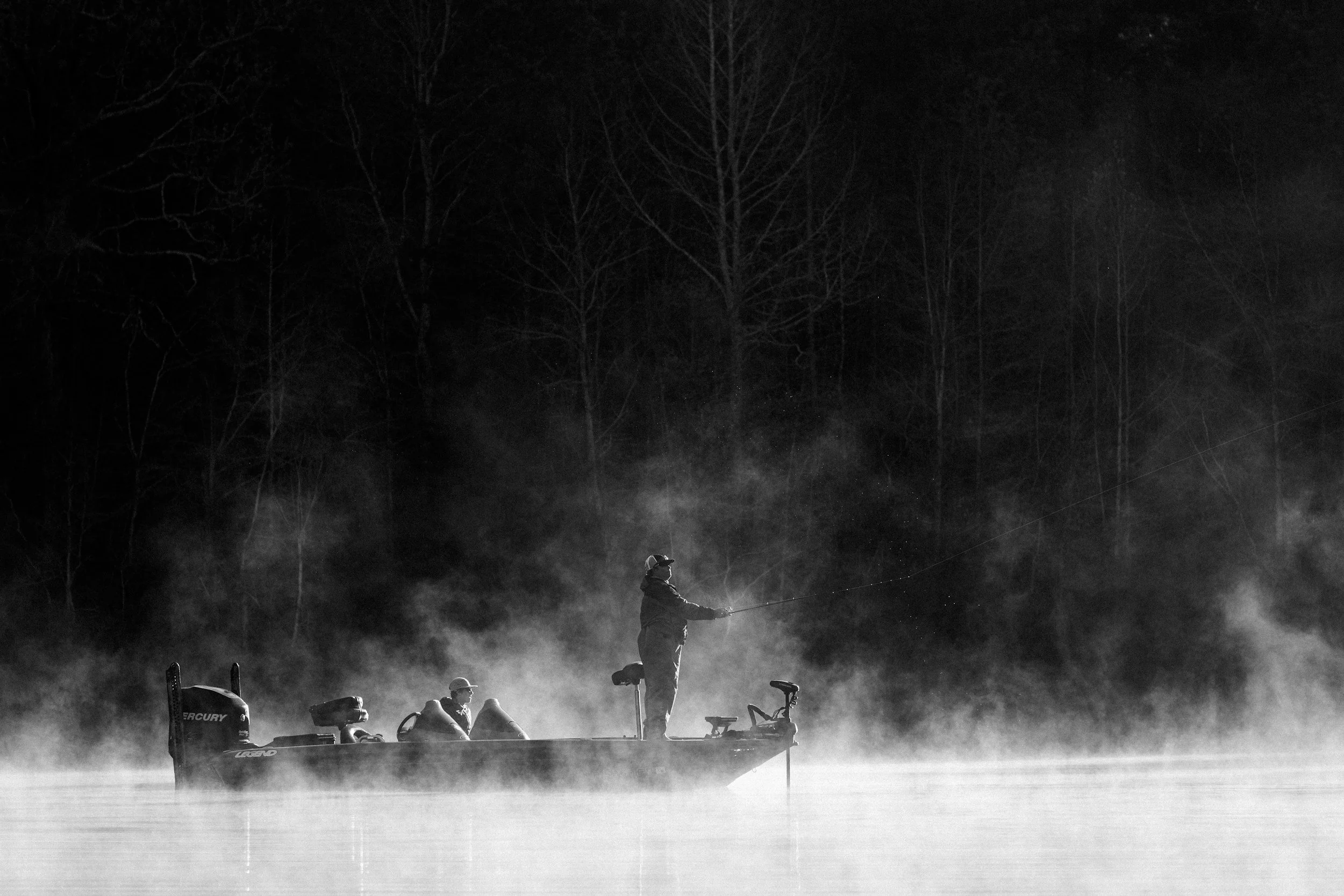 A person fishing from a boat on a foggy lake at night, with three other individuals on the boat, and a backdrop of leafless trees.