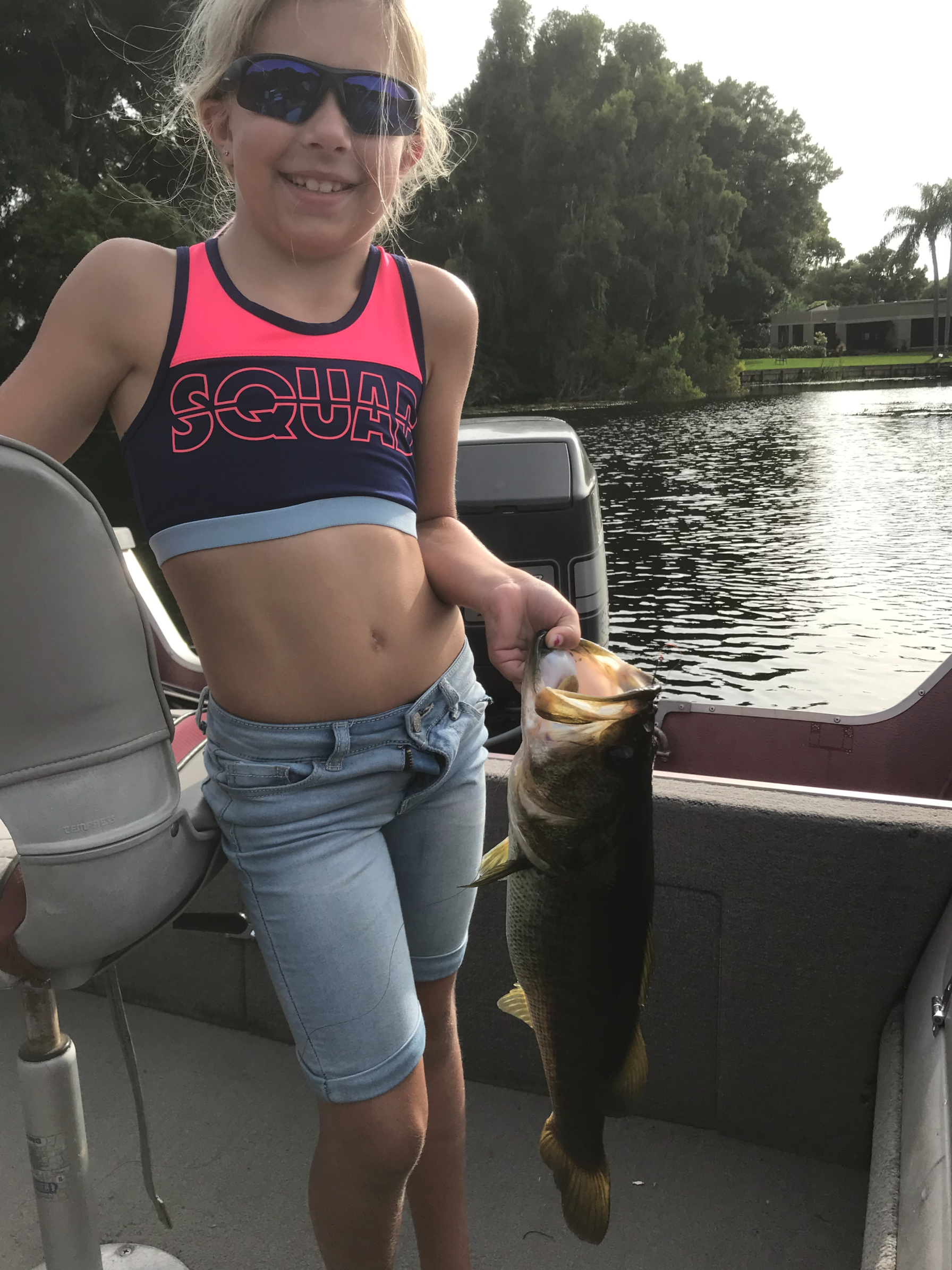 4lb Bass A young girl in sunglasses, a sporty tank top, and denim shorts is on a boat holding a large fish she caught, with water and trees in the background.