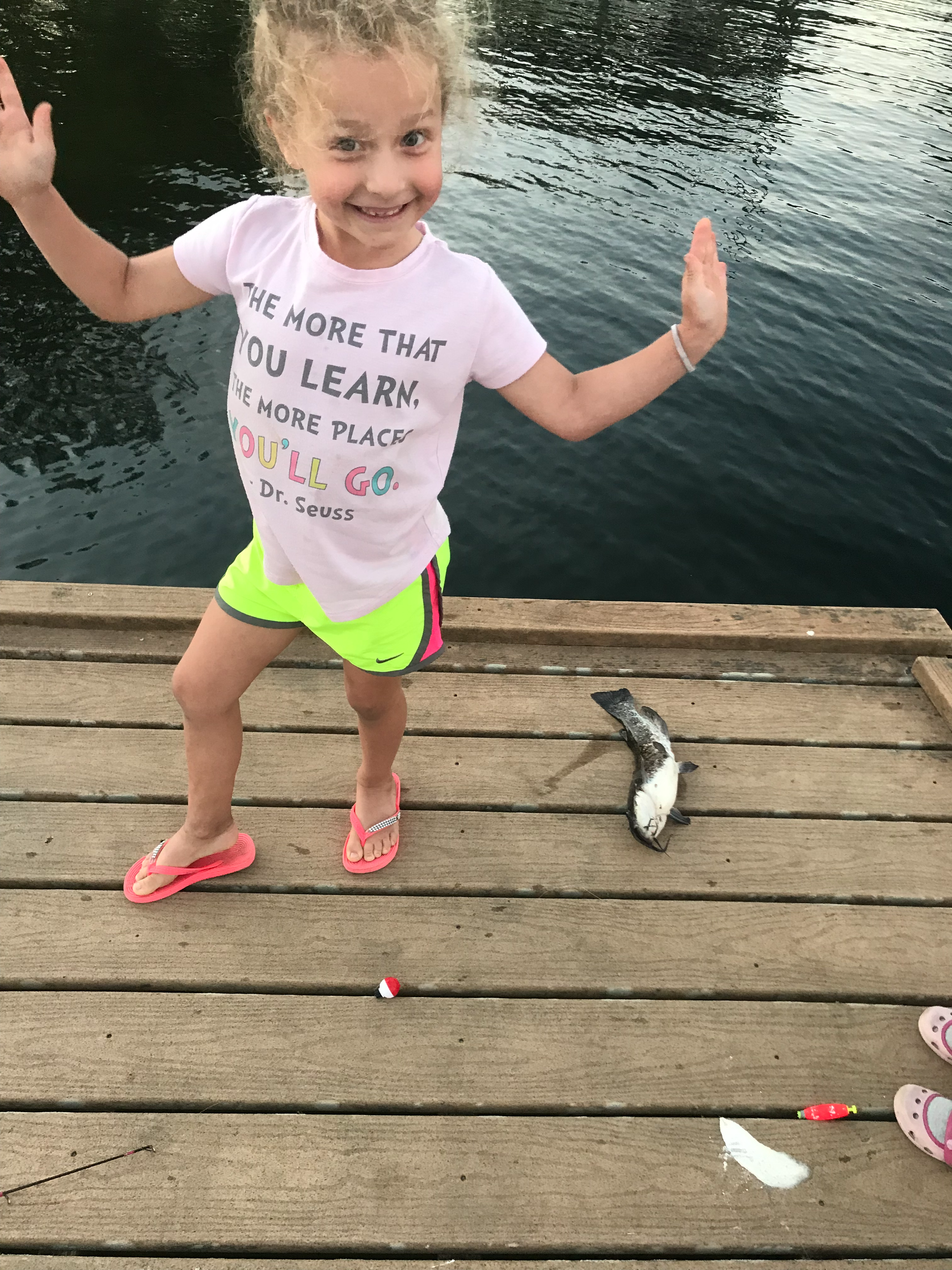 1st Catfish A young girl standing on a wooden dock by a body of water, smiling, with a fish lying on the dock near her feet, and fishing gear on the dock.