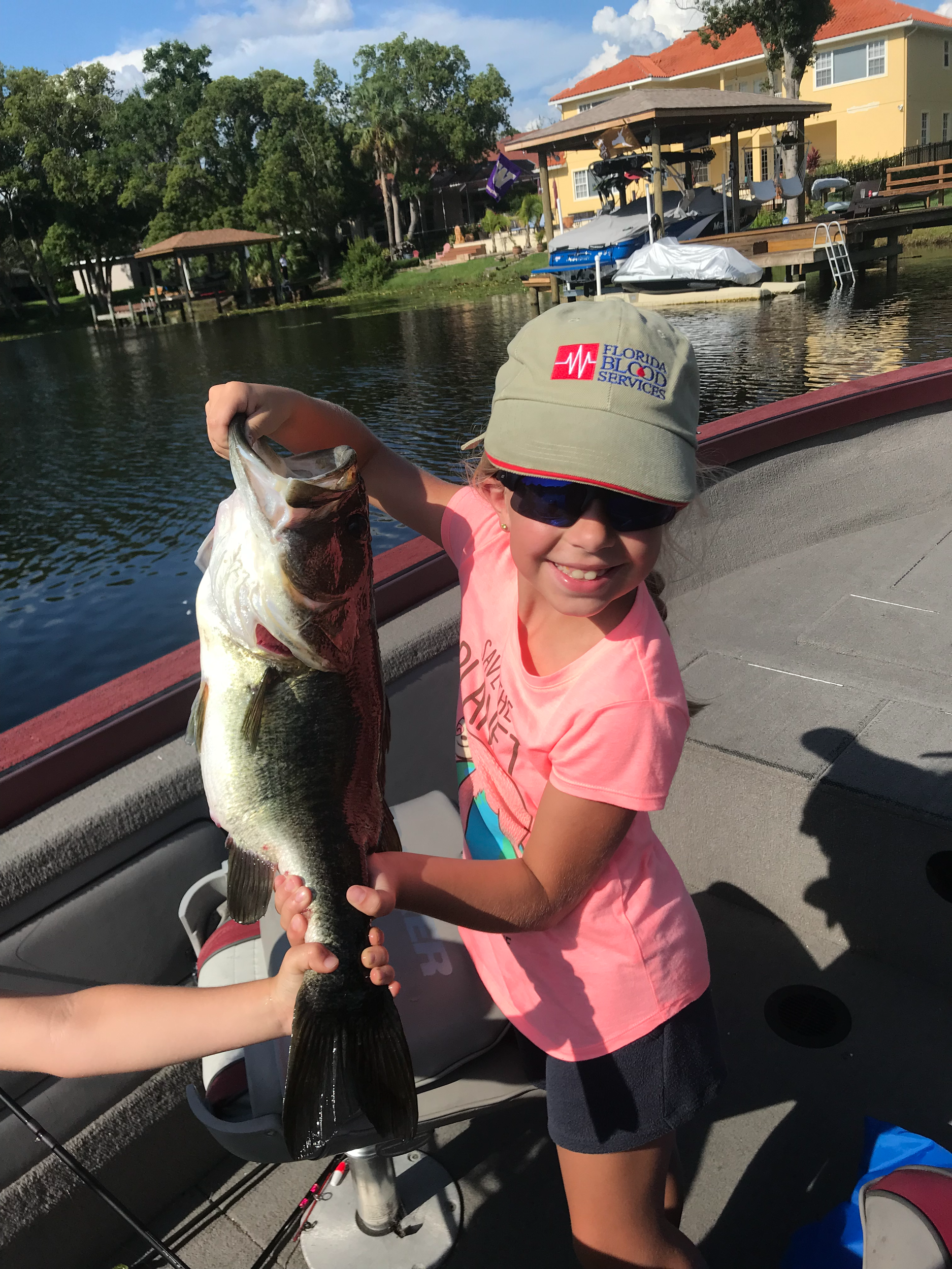 7lb Bass Young girl smiling and holding a large bass fish on a boat by a lake with houses and trees in the background.