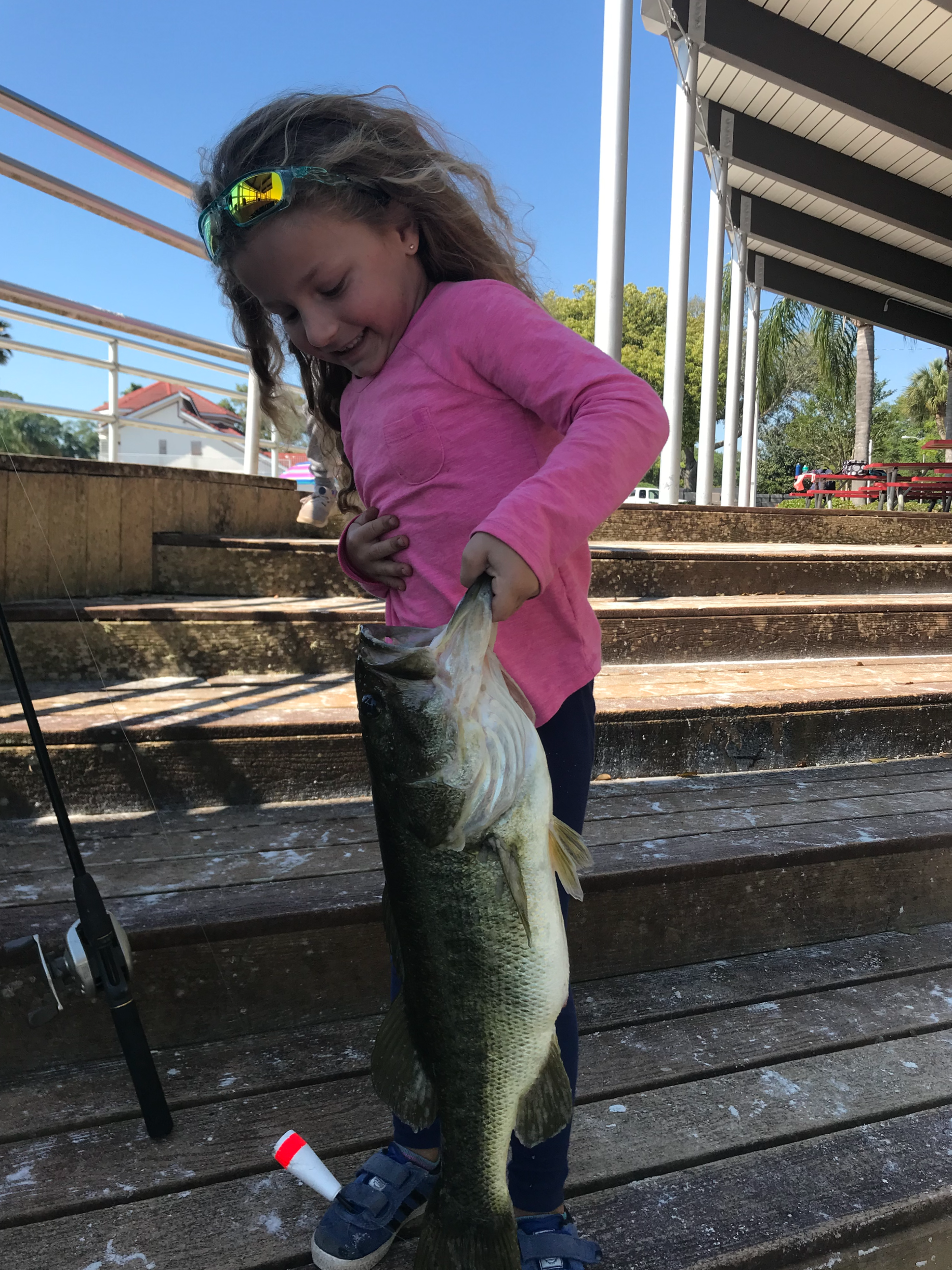6.6 lb Bass Young girl with sunglasses on her head, smiling, holding a large fish she caught by the mouth on a wooden dock outdoors.