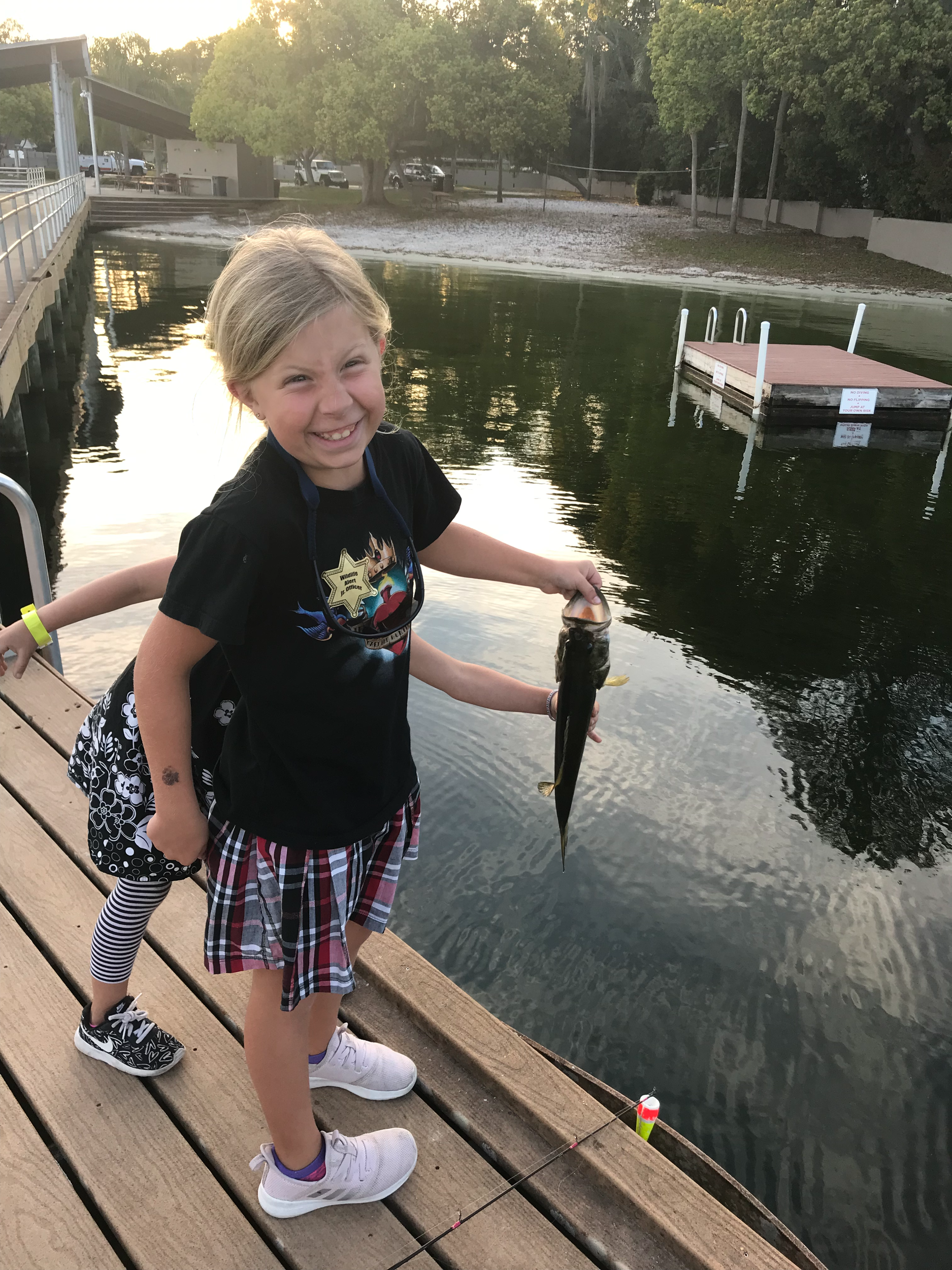 A young girl with a big smile holding a fish she caught, standing on a wooden dock near a body of water, with a fishing pole in the water and trees in the background.