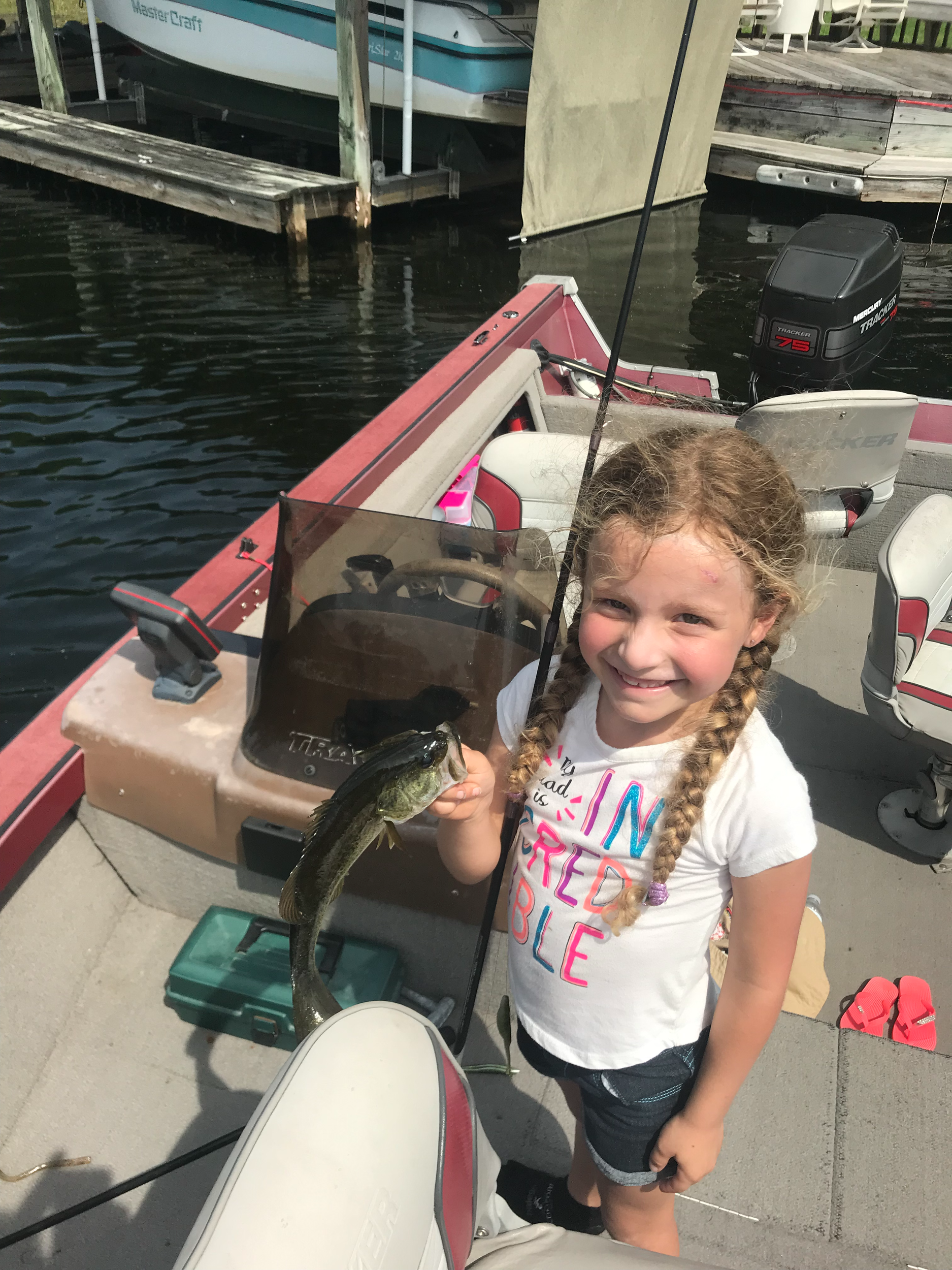 1st Bass with worm A young girl with braided hair smiling on a boat, holding a fish she caught, with water and other boats in the background.