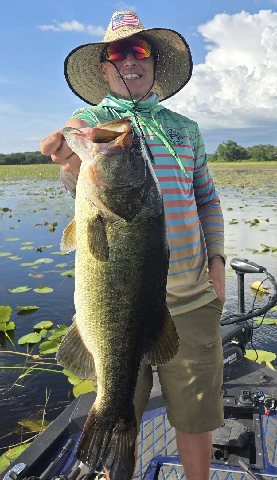 10lb Bass Man wearing a wide-brimmed hat, sunglasses, and a striped long-sleeve shirt holding a large fish while standing on a boat in a water body with lily pads, with a partly cloudy sky in the background.