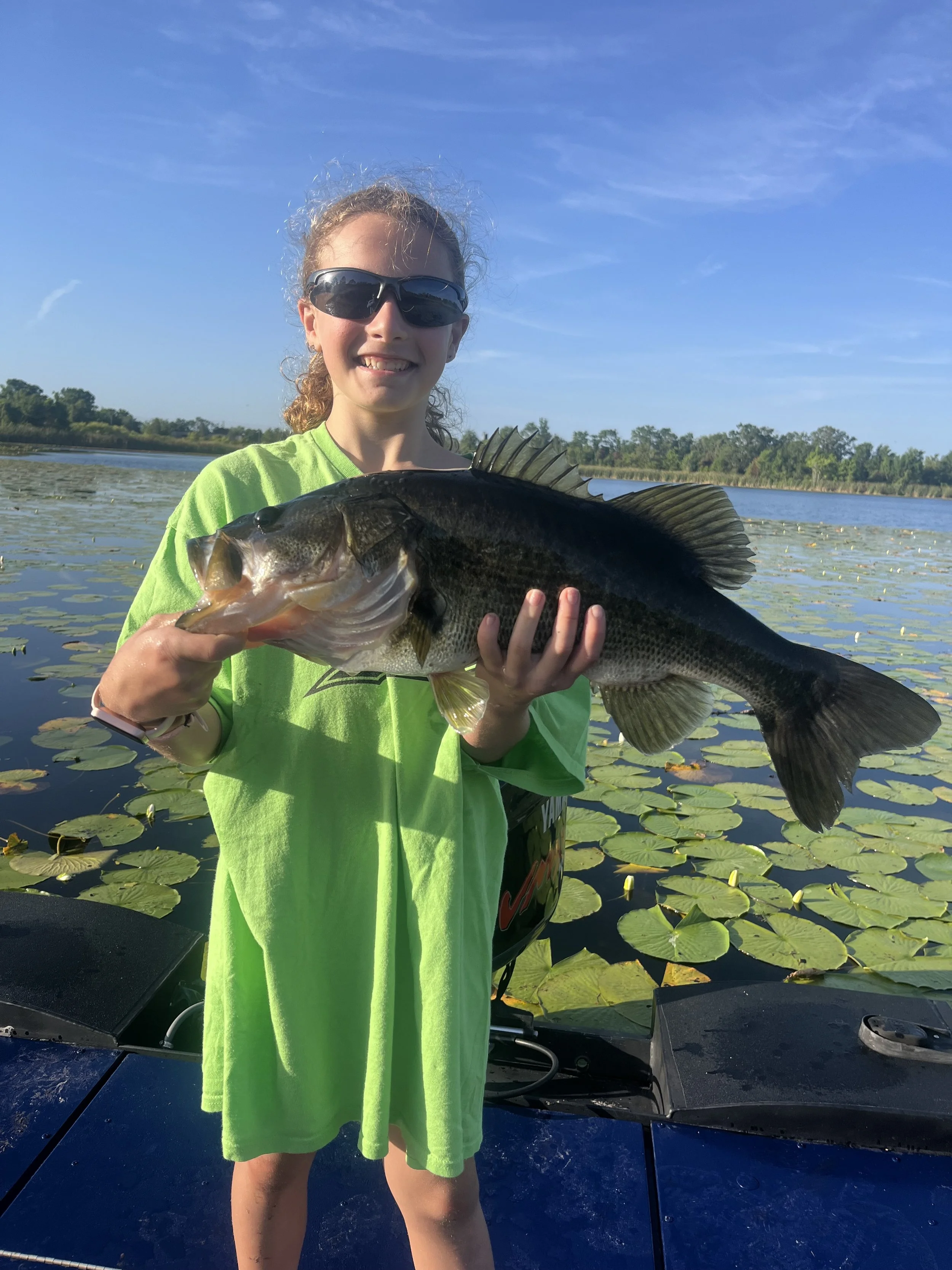 6lb Bass A young girl in a green shirt and sunglasses holding a large fish, standing on a boat surrounded by water lilies and water with trees in the background.
