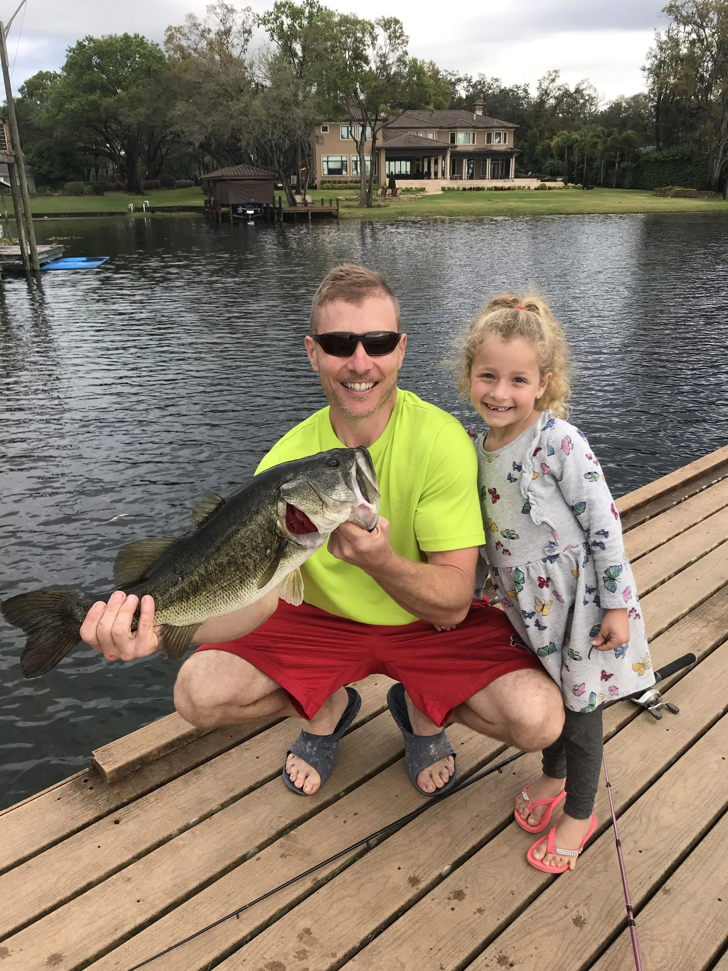 8lb Bass A man and a young girl on a wooden dock by a lake. The man is holding a large fish and smiling, while the girl is standing next to him with a happy expression. There are houses and trees in the background.